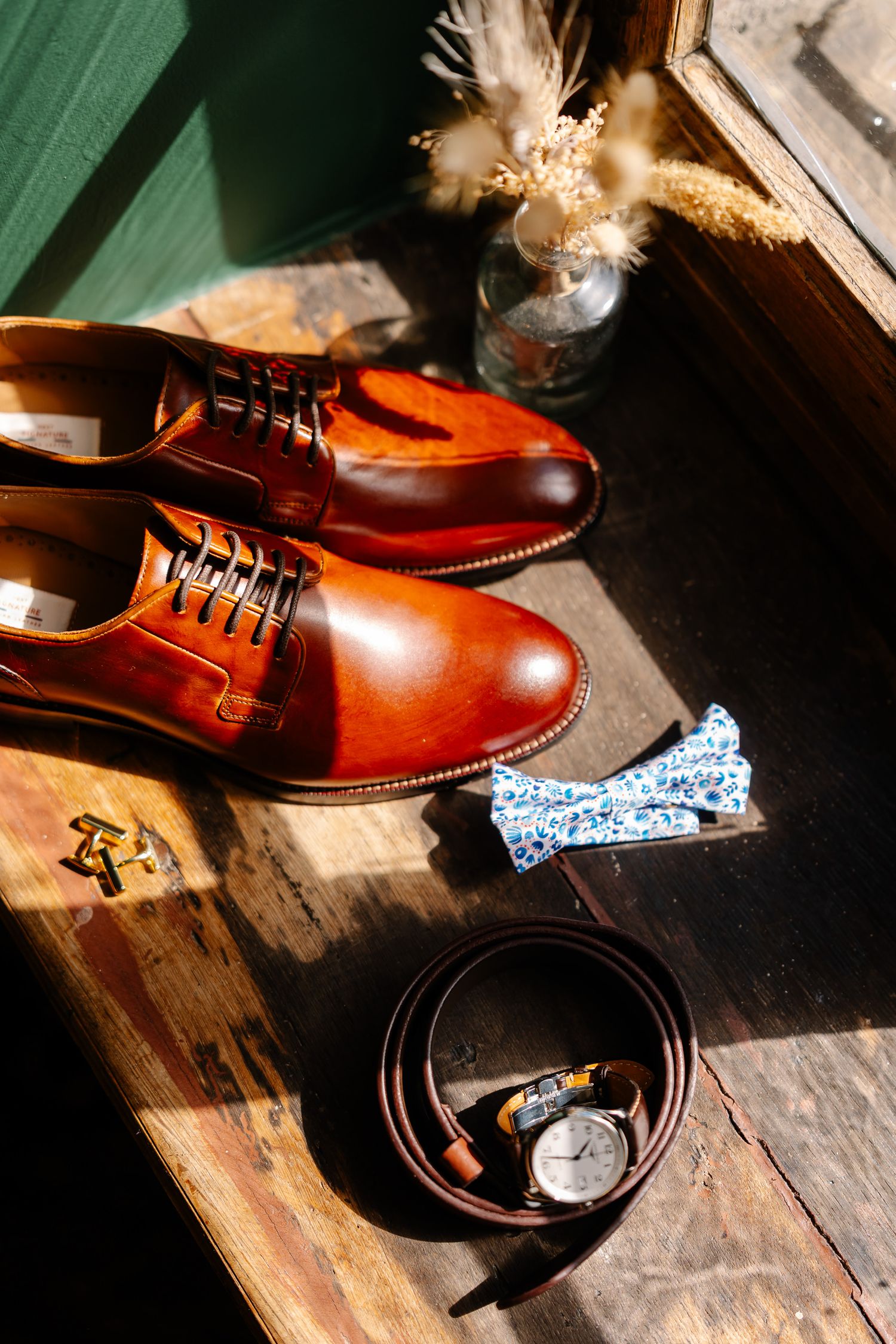 Brown leather dress shoes, floral bow tie, leather belt and watch arranged on rustic wooden surface in natural lighting.