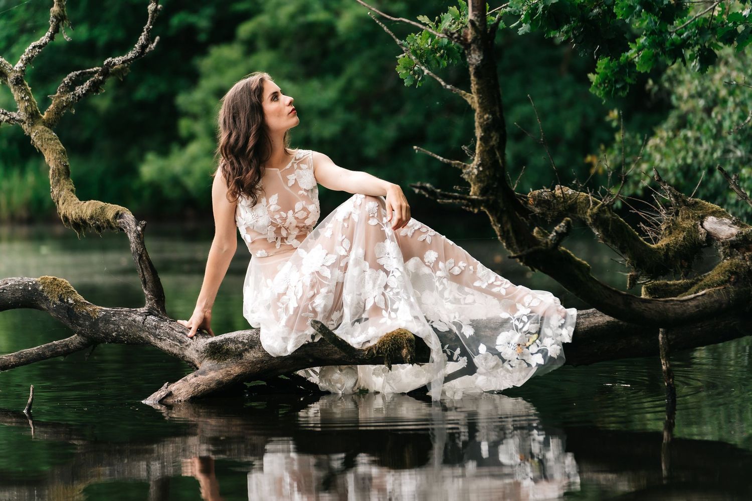 A bride in a white lace dress sits gracefully on a fallen tree branch over a serene pond.