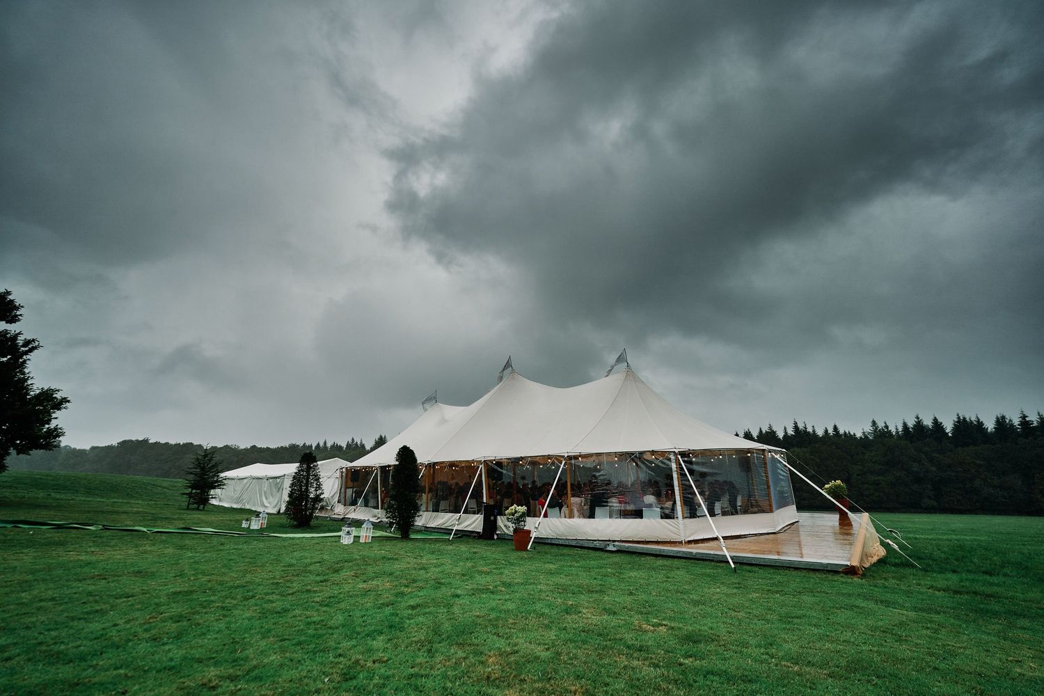Dramatic stormy clouds loom over a green field with white event tents set up for an outdoor gathering.