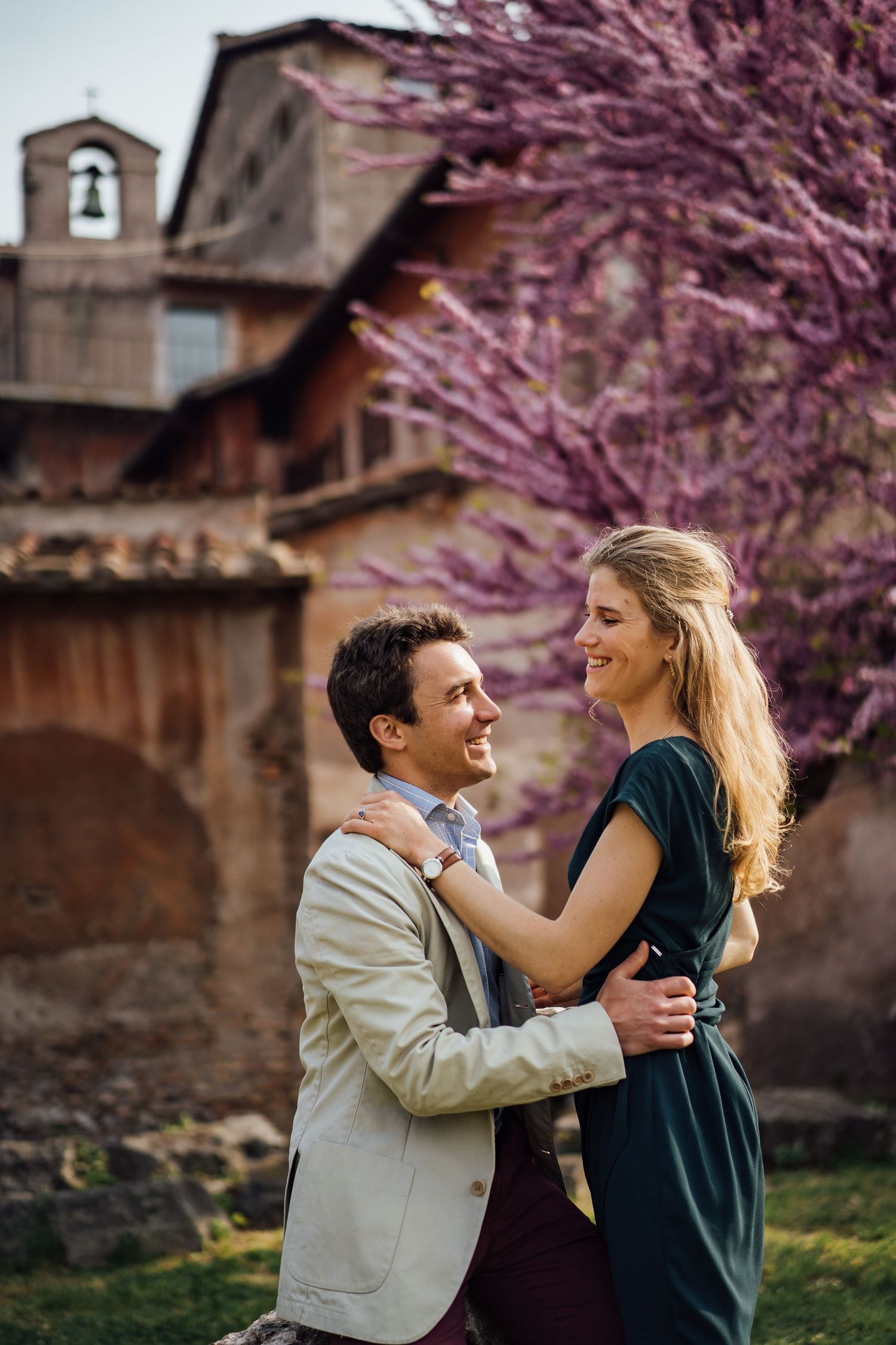 A couple embraces beneath blooming cherry blossom trees against a rustic stone building backdrop.