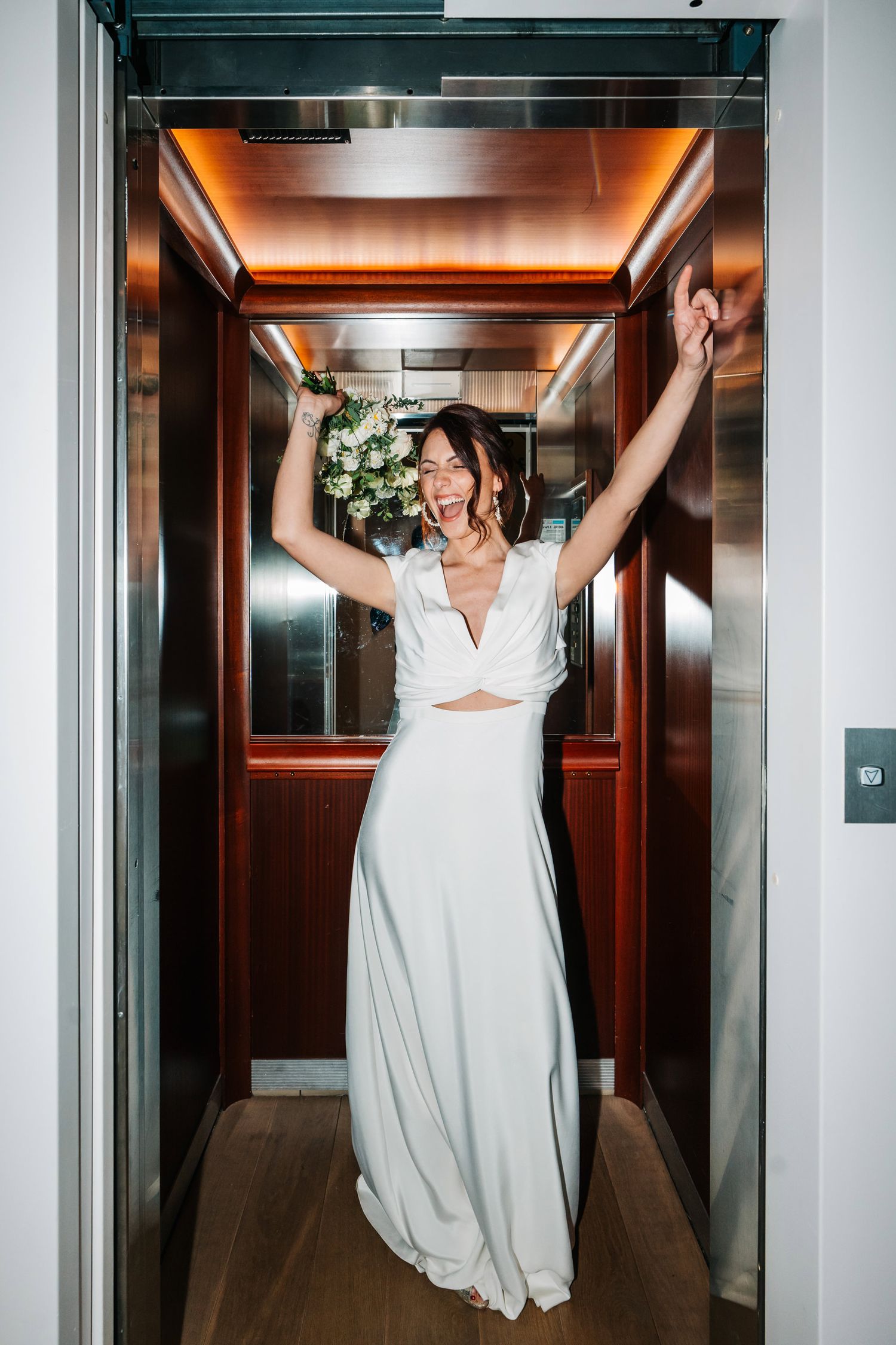 A bride in a white wedding dress poses playfully in a modern elevator, creating a fun series of shots.