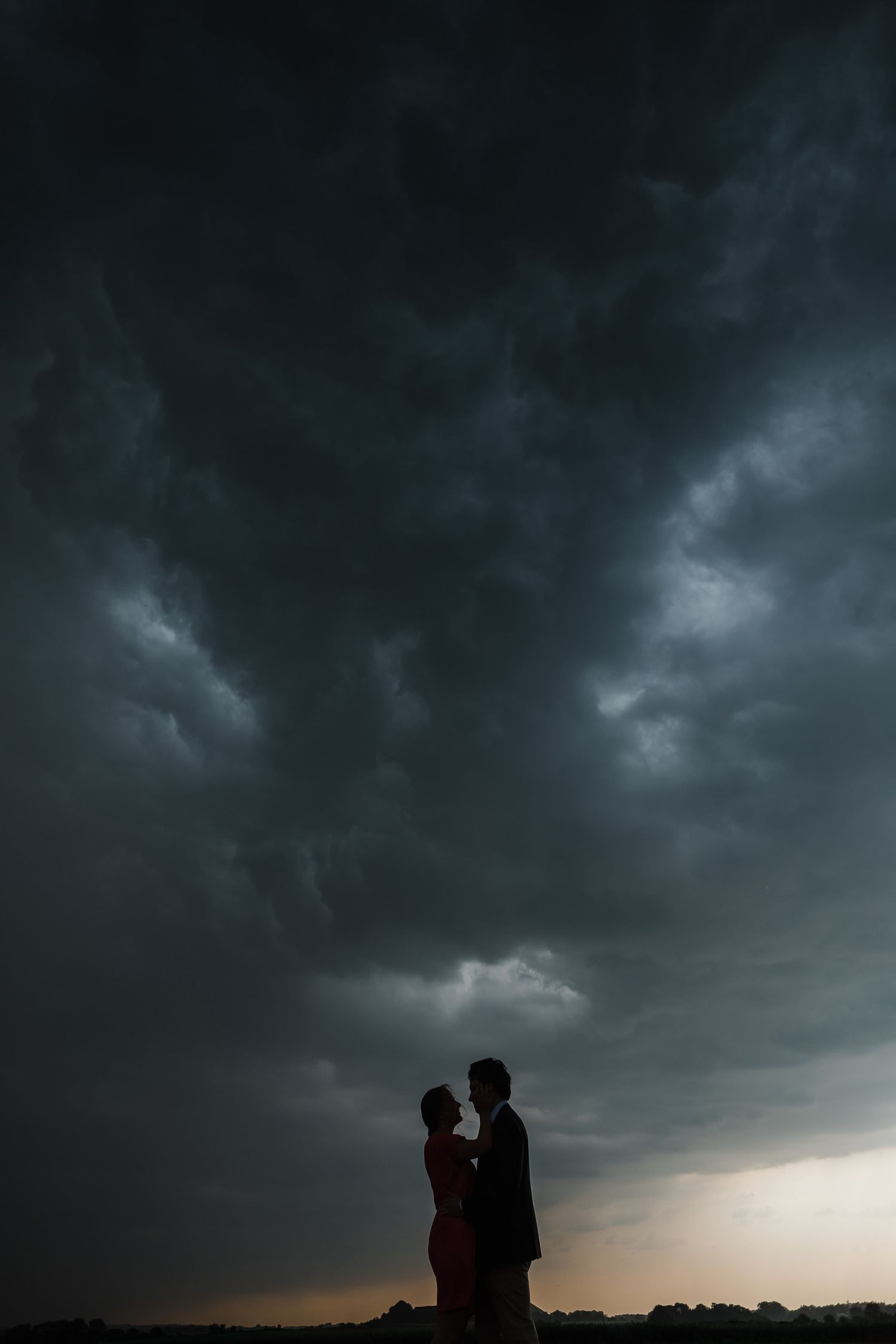 Dark stormy clouds loom over silhouetted figures standing together against a dramatic sunset sky.