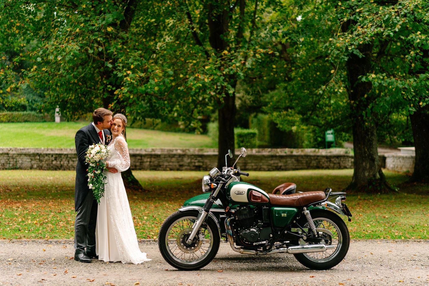 Romantic wedding couple poses with vintage motorcycle in lush green park setting.