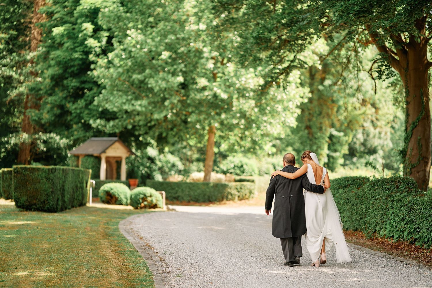 Couple walking arm in arm down a garden path surrounded by manicured hedges and trees.