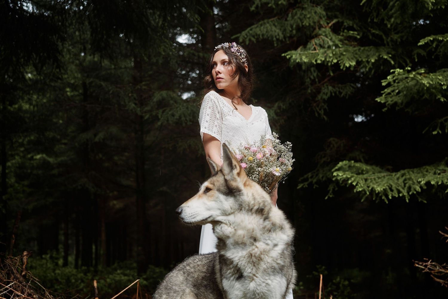 A woman in a white dress sits with a large wolf-like dog on a wooden dock by a serene lake.