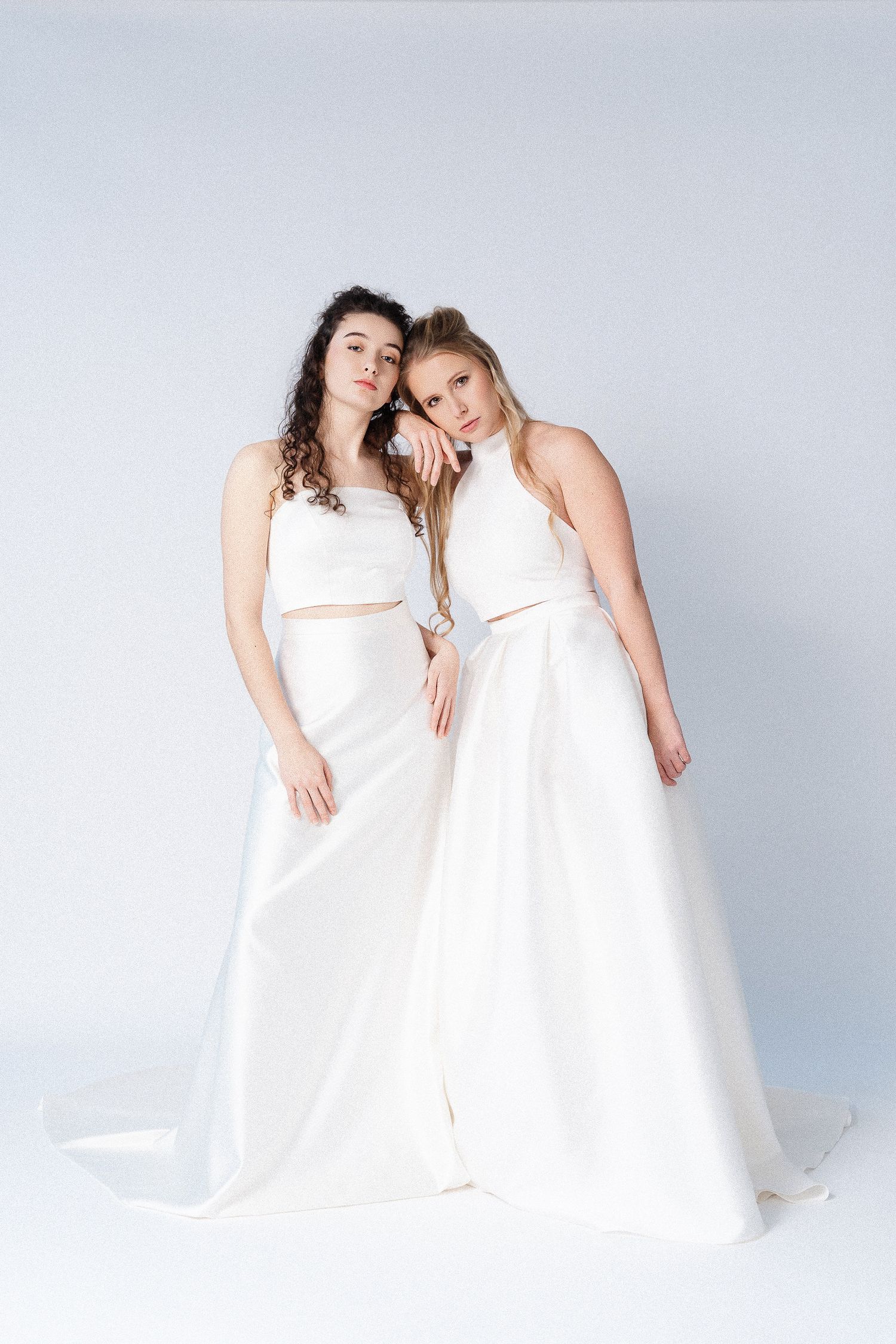 Two figures in flowing white dresses pose together against a light background in an artistic fashion photoshoot.