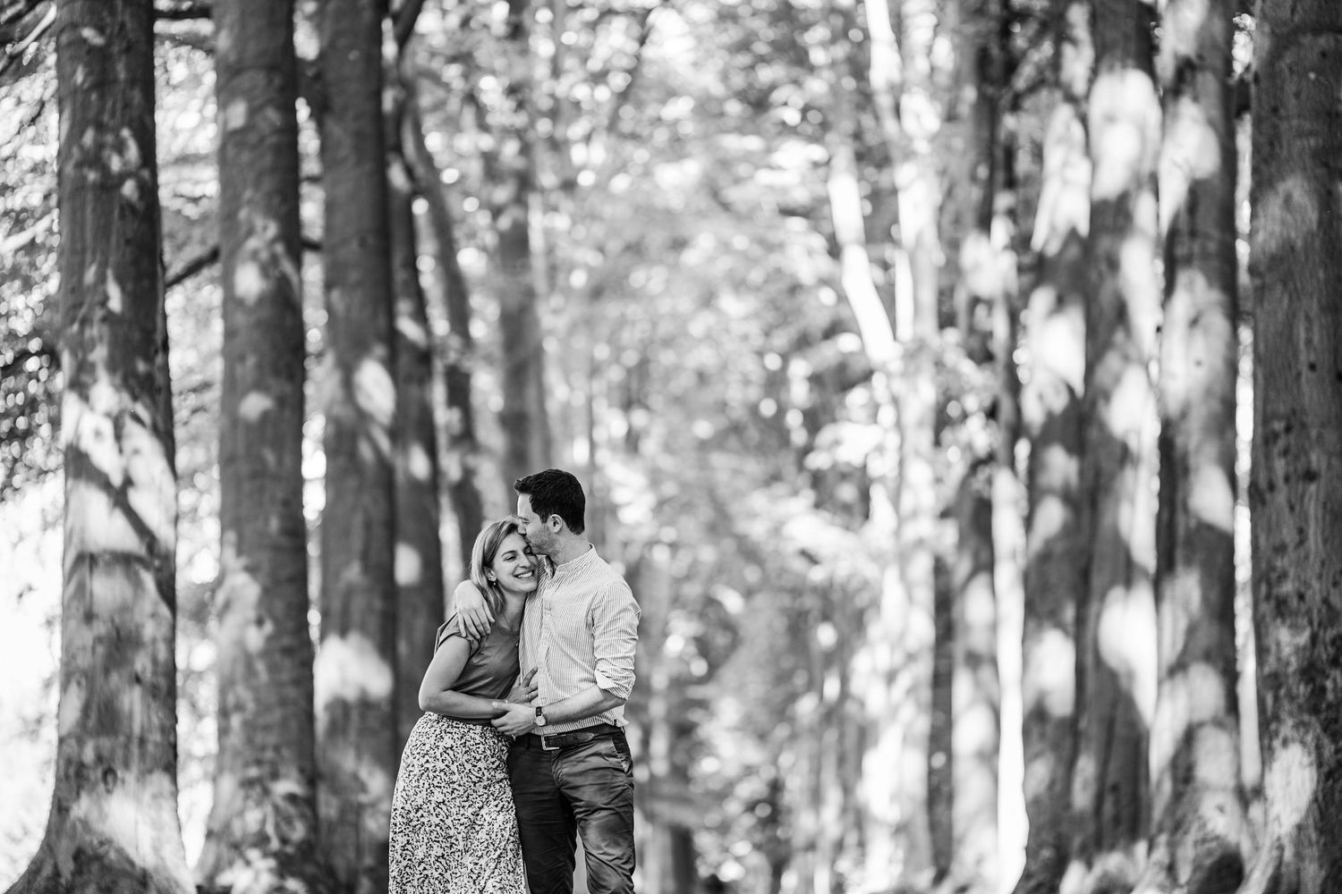 A black and white photograph shows a couple in a wooded forest setting sharing a tender moment together.