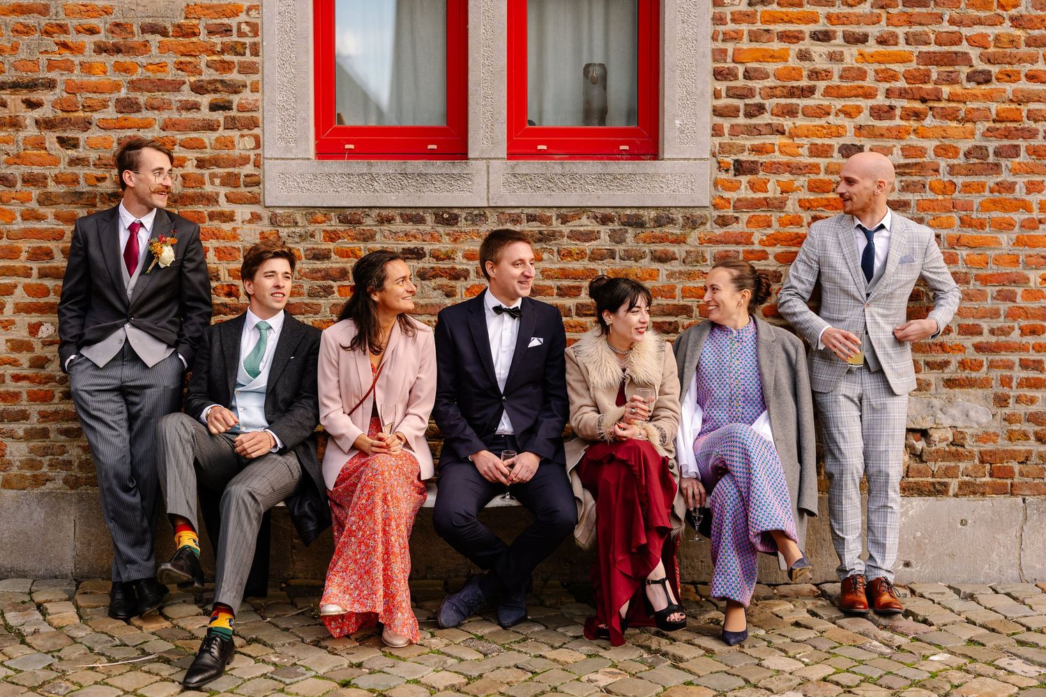 Wedding party sits casually against rustic brick wall while showing off colorful socks in coordinated pose.