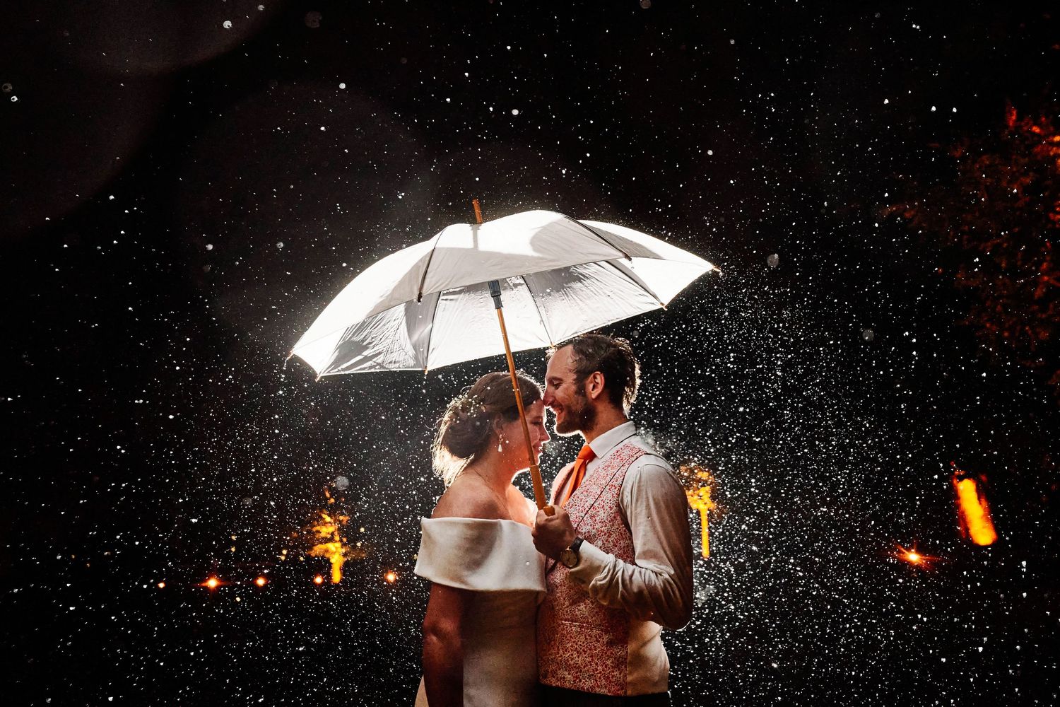 Couple stands under a white umbrella in the rain at night with glowing lights in the background.