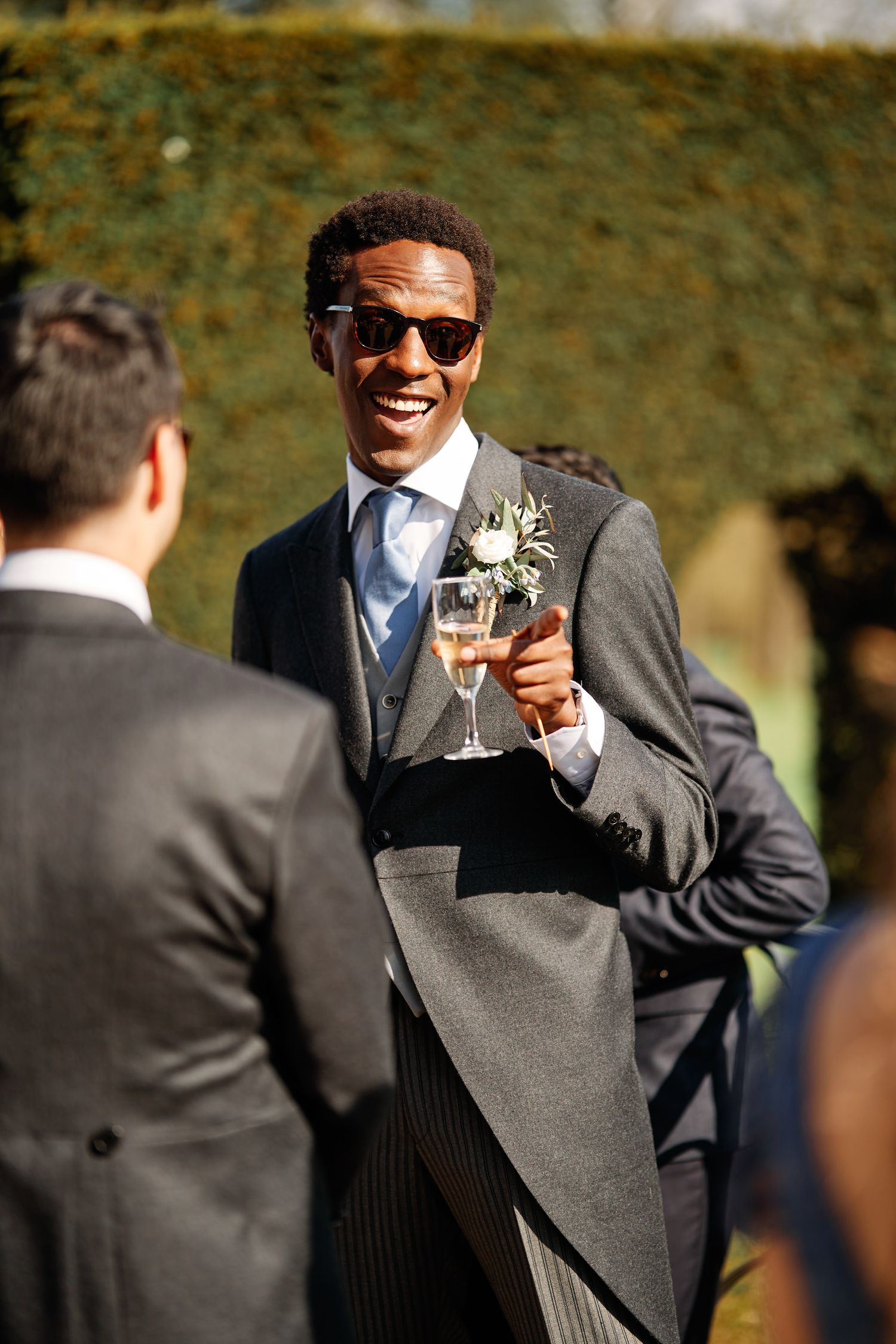 Wedding guest in a grey suit and sunglasses raises a glass in celebration during an outdoor reception.