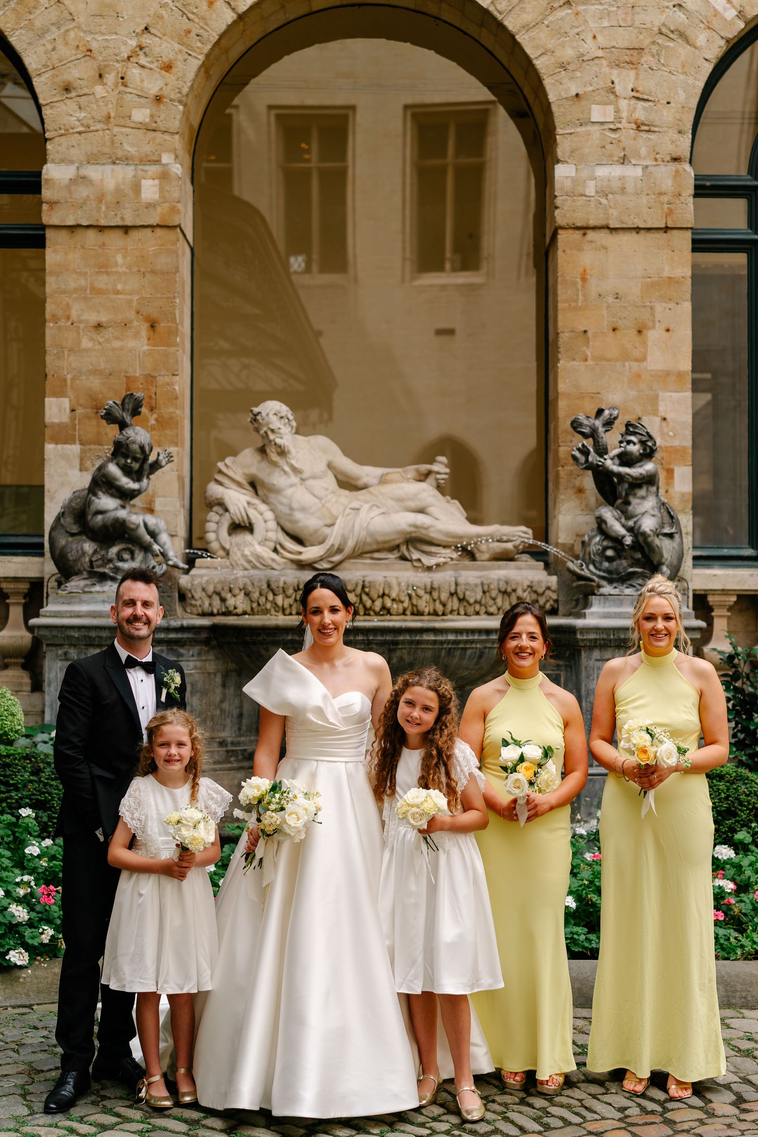 Wedding party poses in front of stone archways with classical sculptures and white flowers in a courtyard.