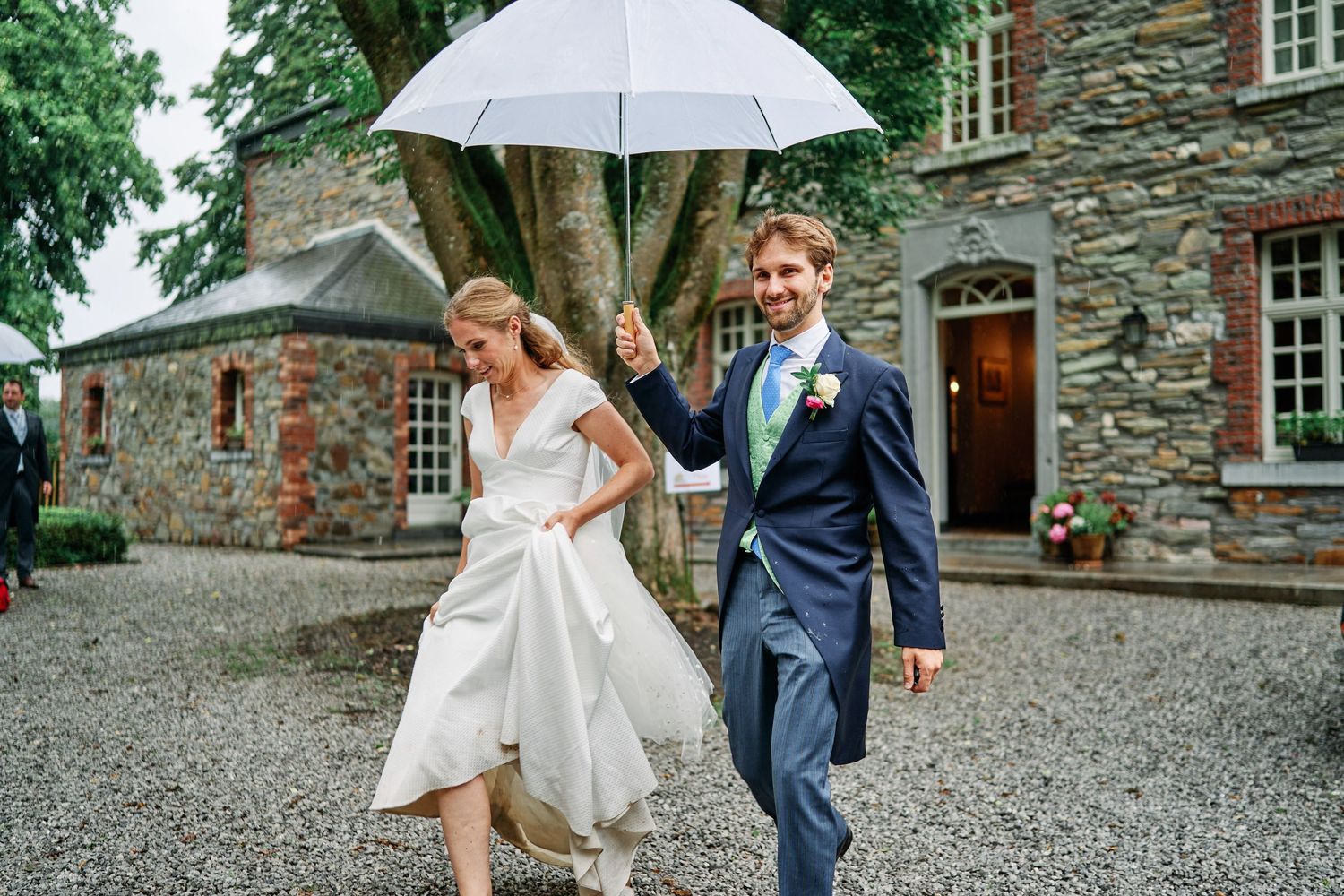 A couple walks together under a white umbrella on a rainy day outside a rustic stone building.