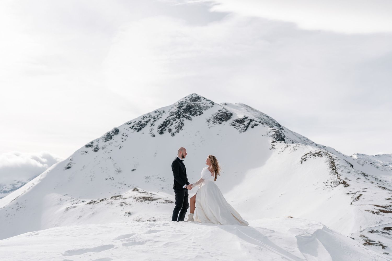 Two people stand together on a snow-covered mountain peak with dramatic white winter landscape in the background.