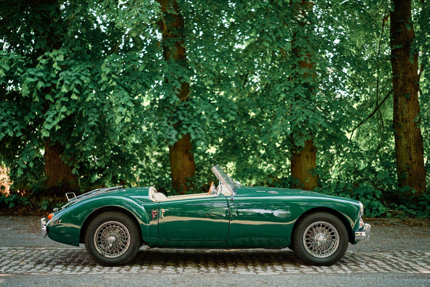 A classic green MG sports car parked in front of lush green foliage on a cobblestone street.
