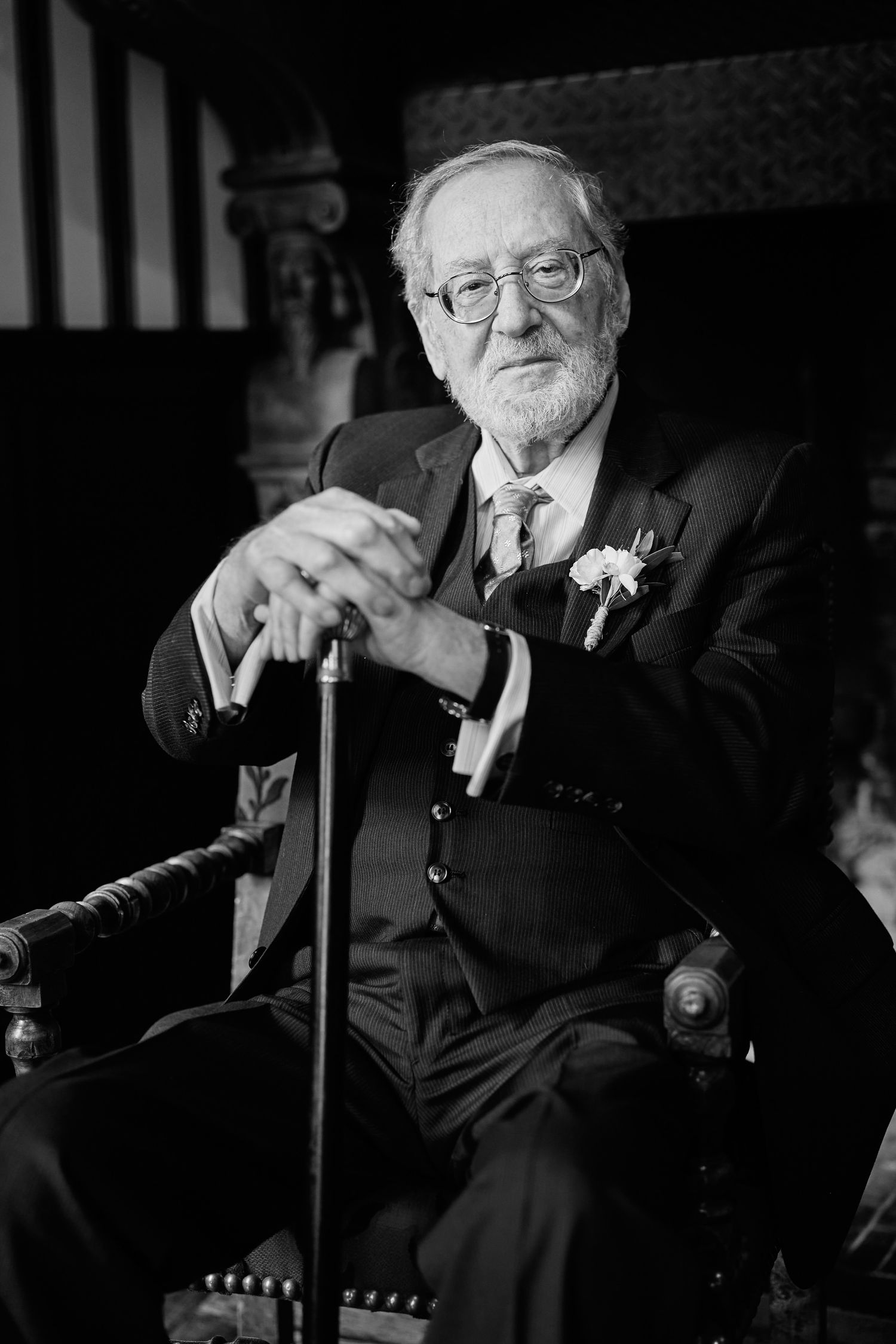 Black and white portrait of an elderly person in formal attire seated with a cane and wearing glasses.