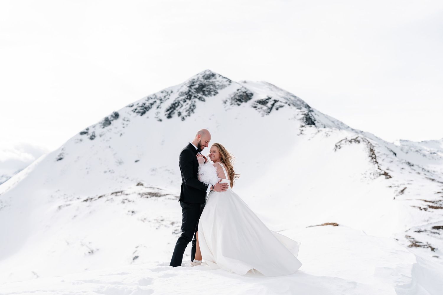 A couple shares a romantic embrace in a snowy winter mountain landscape with dramatic white peaks in the background.