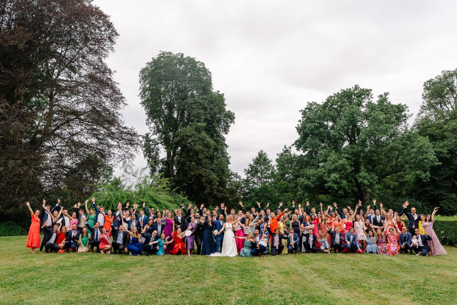 Large wedding party in colorful attire poses together on a green lawn with trees in the background.
