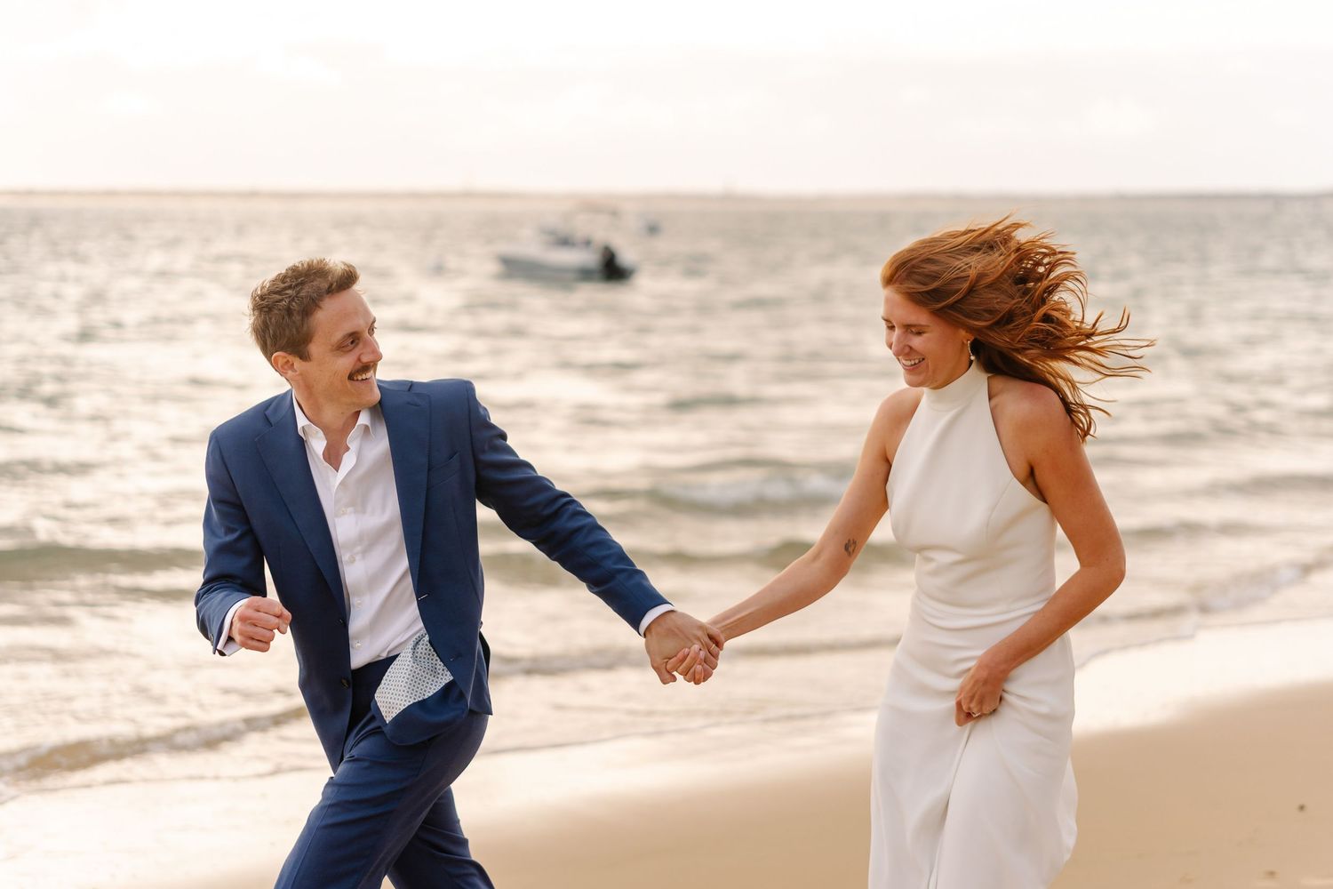 A couple in wedding attire runs joyfully along a beach at sunset, holding hands with the ocean in the background.