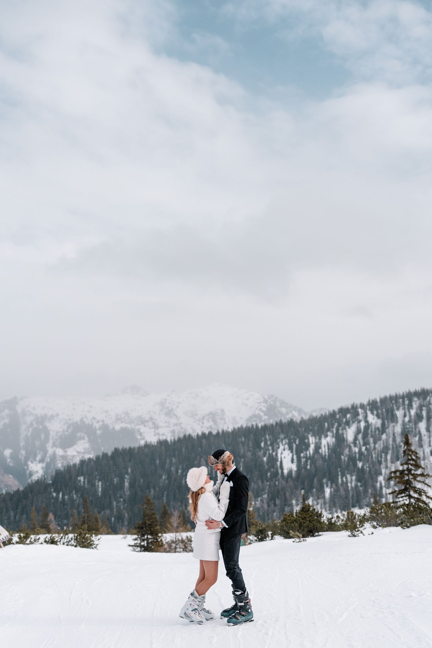 A couple shares a romantic moment in the snow with majestic snow-capped mountains and evergreen forest in the background.