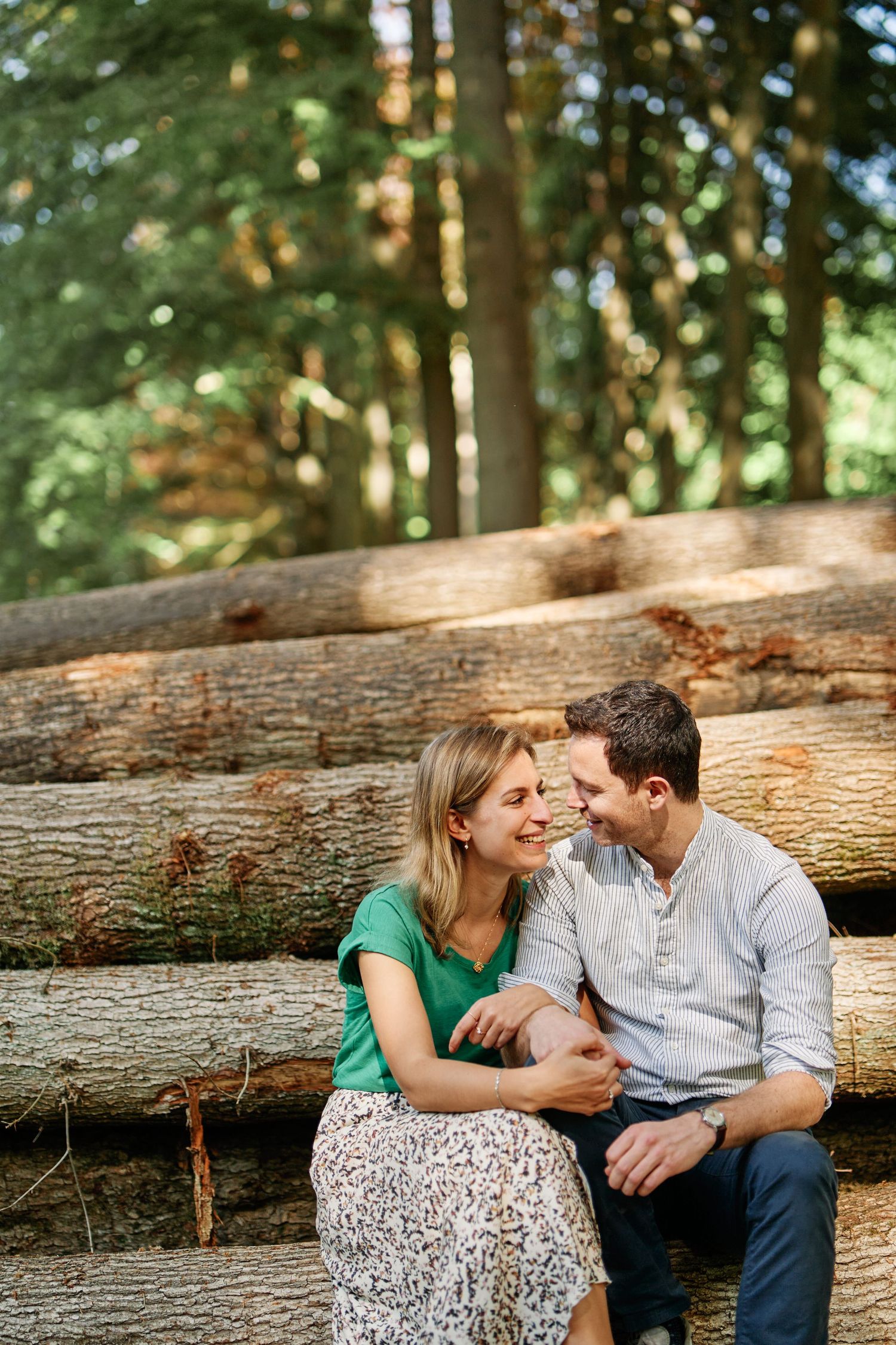 A couple shares an intimate moment while sitting together on fallen logs in a sunlit forest.