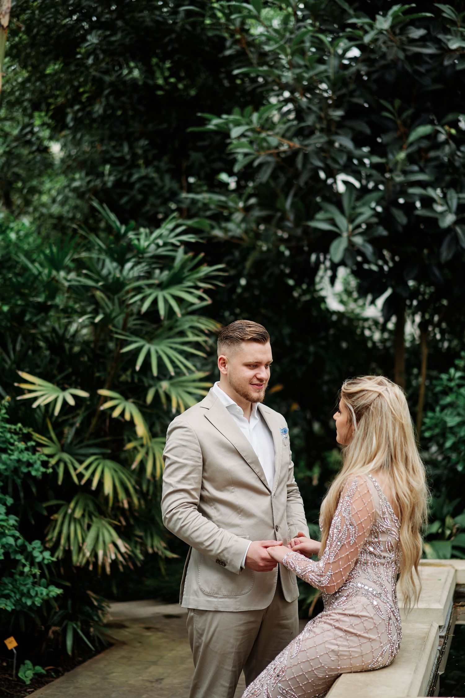 A couple shares a romantic moment in a lush tropical garden setting wearing coordinated beige and blush formal attire.