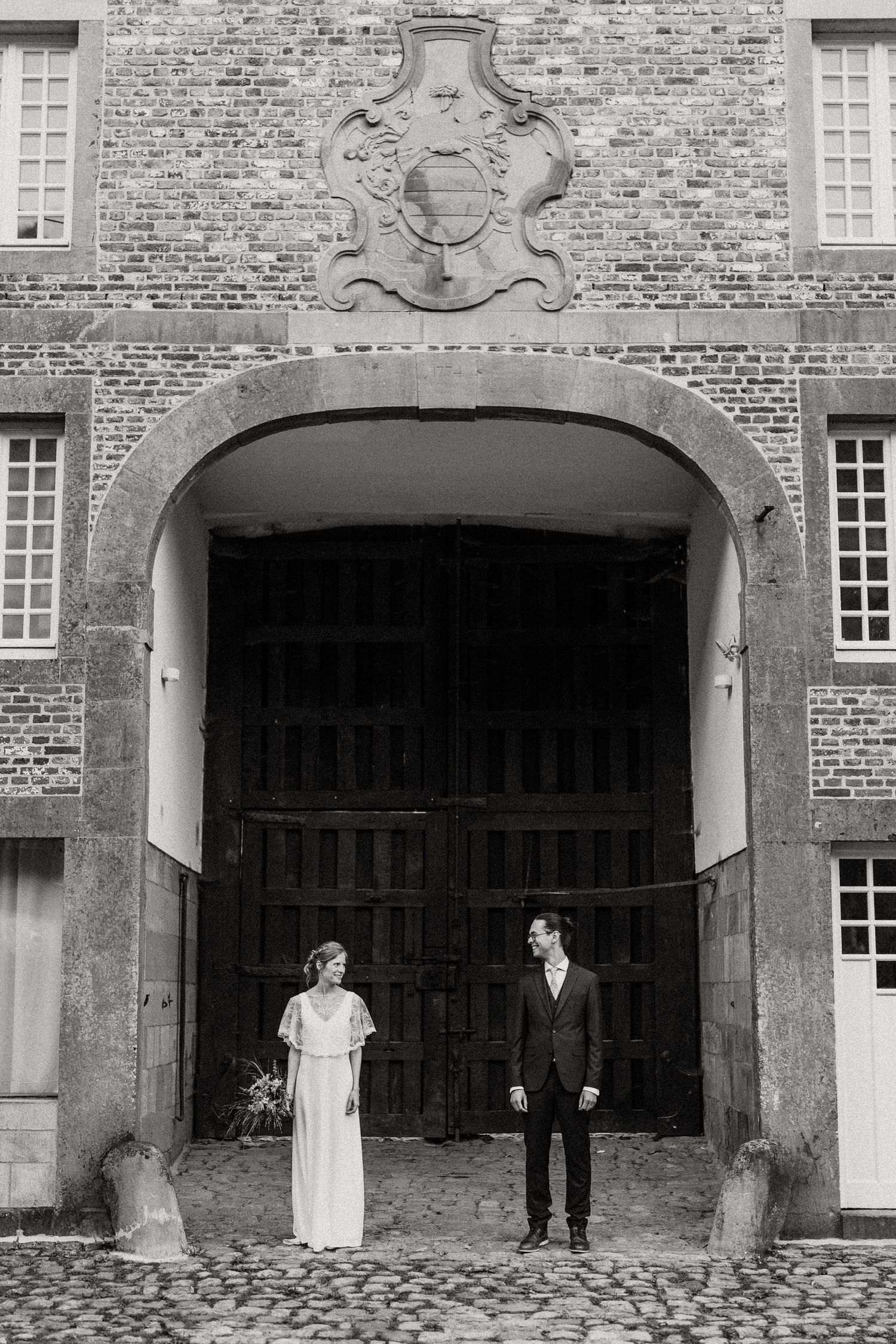 A black and white photograph of two people standing in a grand archway entrance of a historic brick building.