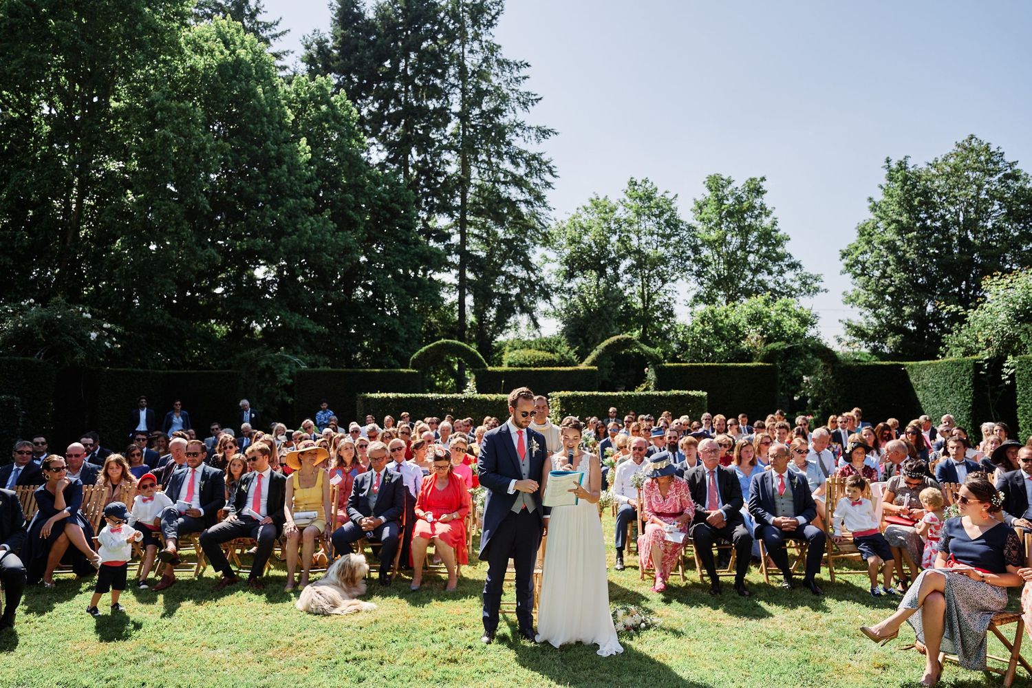 An outdoor summer wedding ceremony with guests seated on the lawn surrounded by lush green trees and gardens.