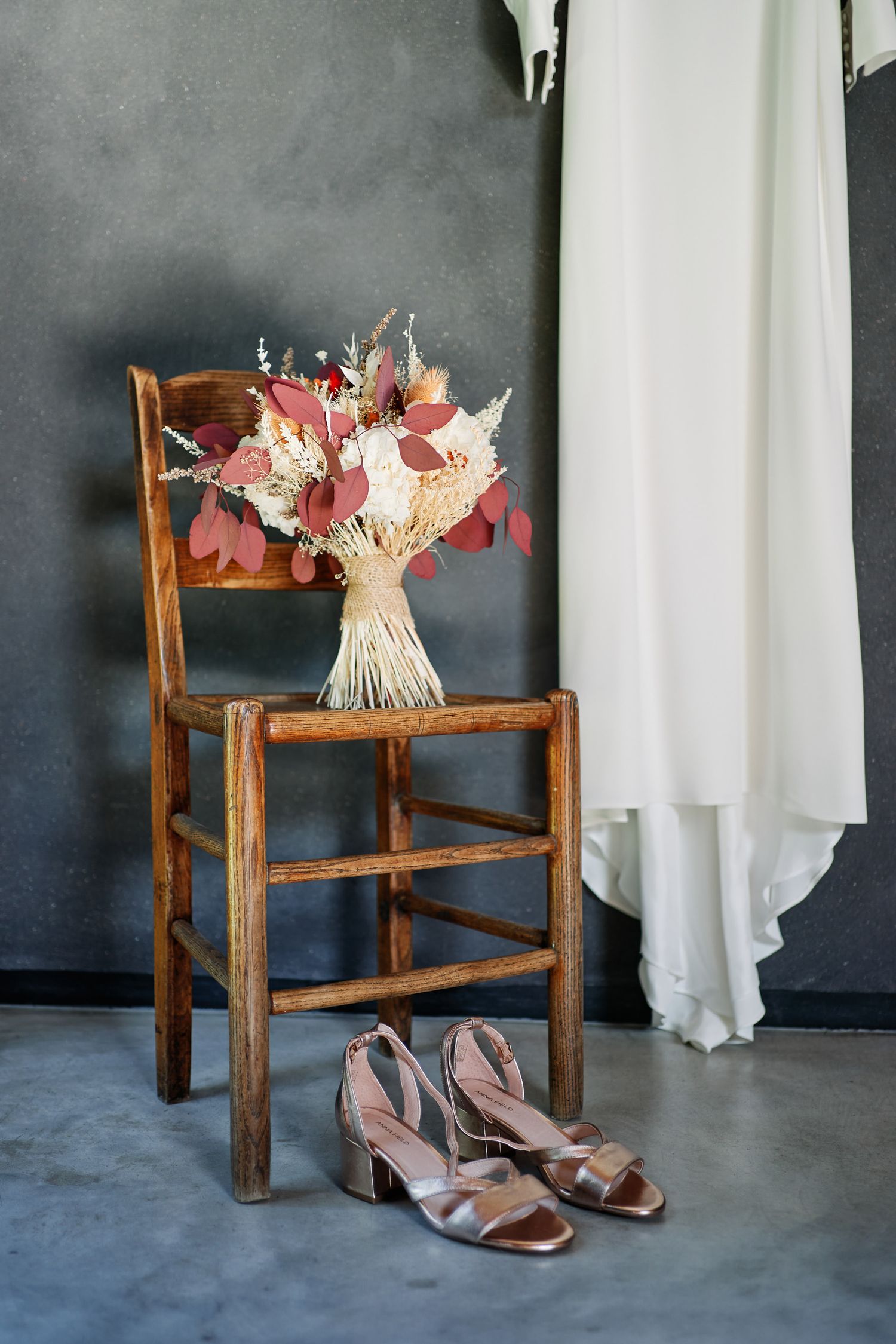 A wedding dress hangs beside a rustic wooden chair with pink floral bouquet and blush heels on concrete floor.