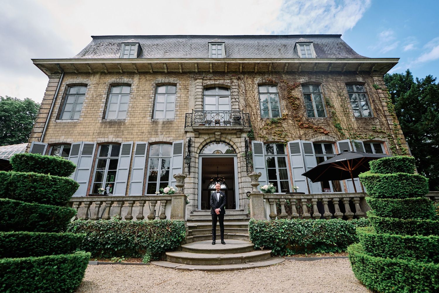 Elegant French-style mansion with symmetrical architecture and manicured topiary gardens leading to grand entrance steps.