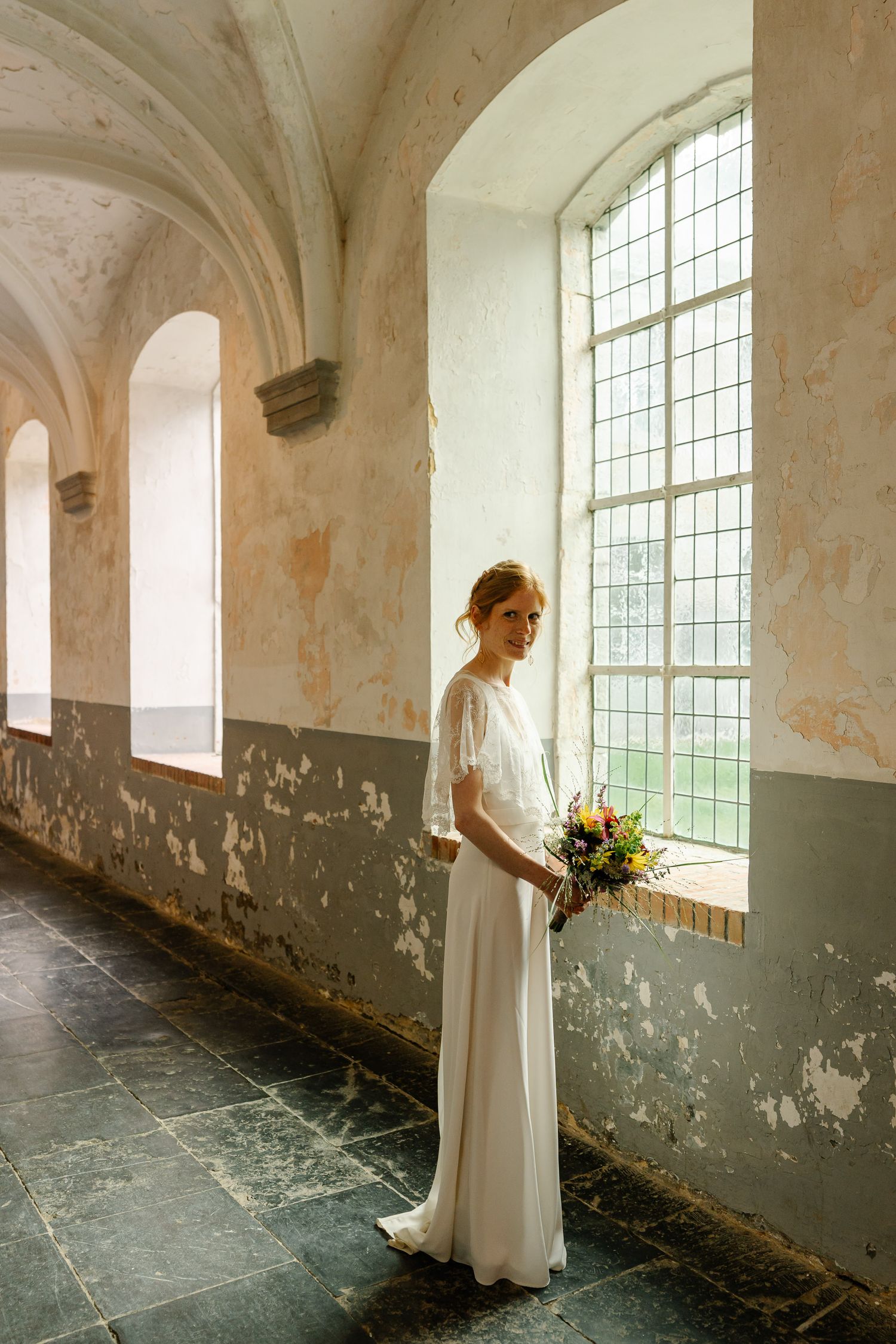 A person in a white dress stands by large windows in an old abandoned building with peeling walls.