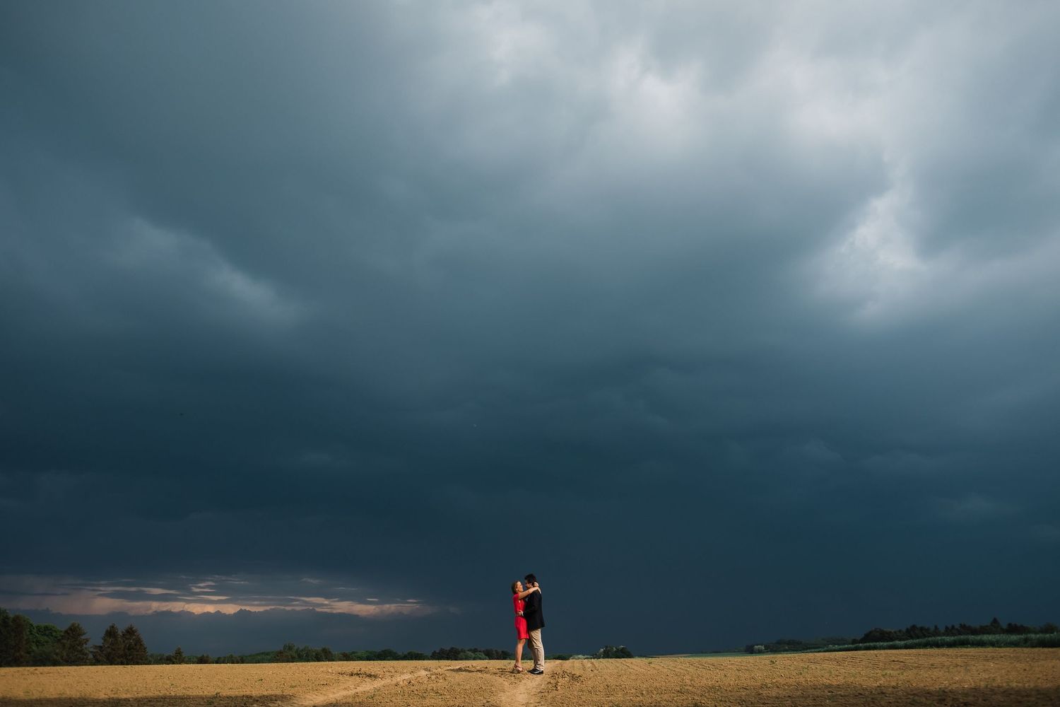 Storm clouds gather over a prairie landscape as a couple stands silhouetted against the darkening horizon.