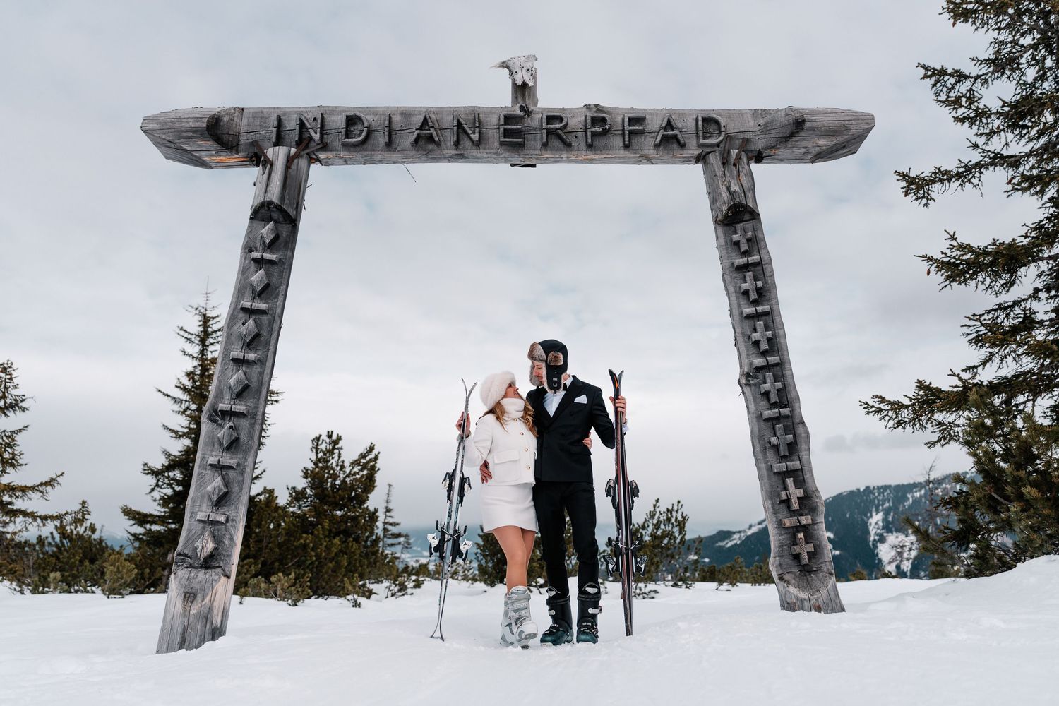 A panoramic view of snowy mountains with a wooden Indian Head sign and people standing near the summit.