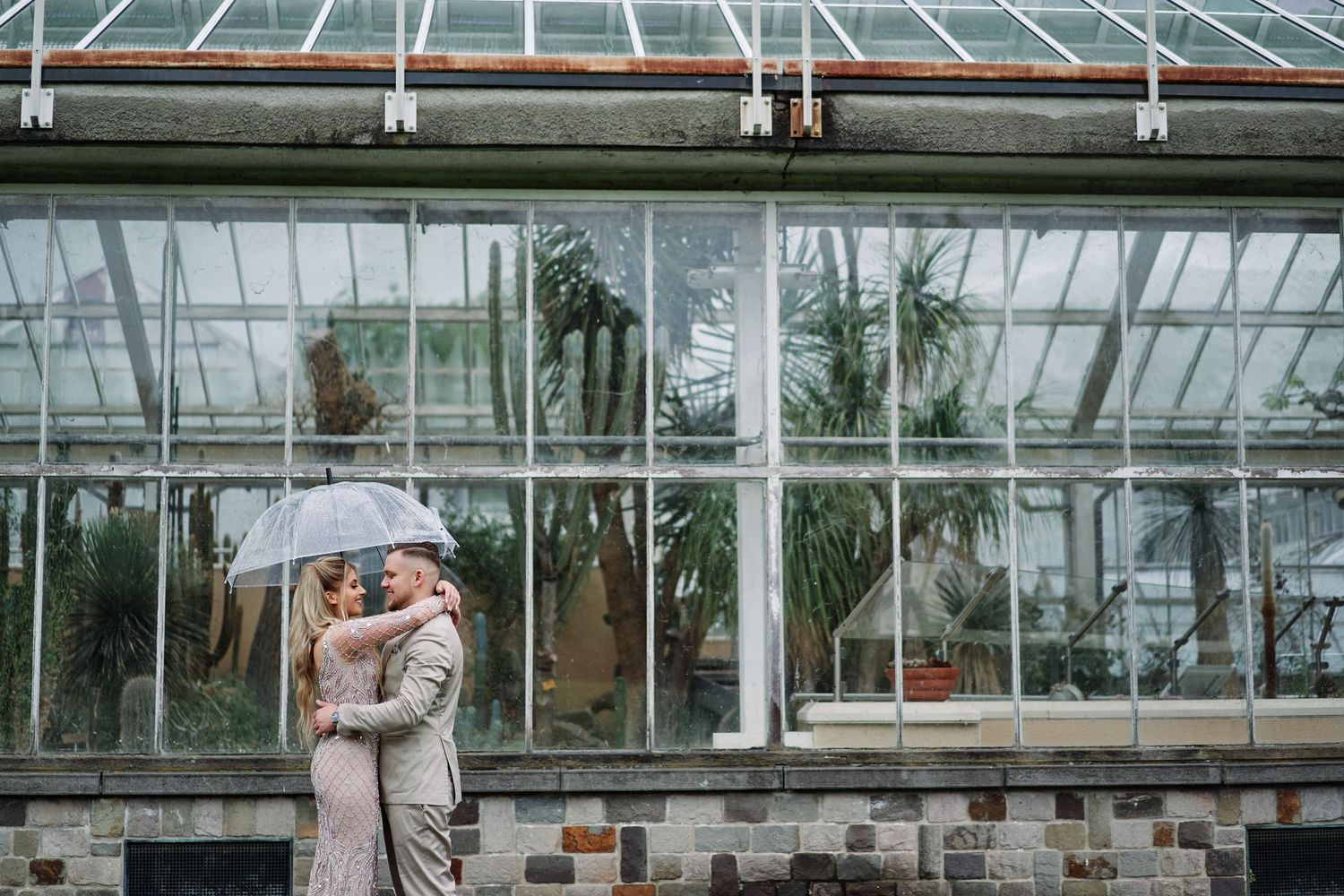 Elegant wedding couple embraces in front of a glass greenhouse conservatory filled with natural light.