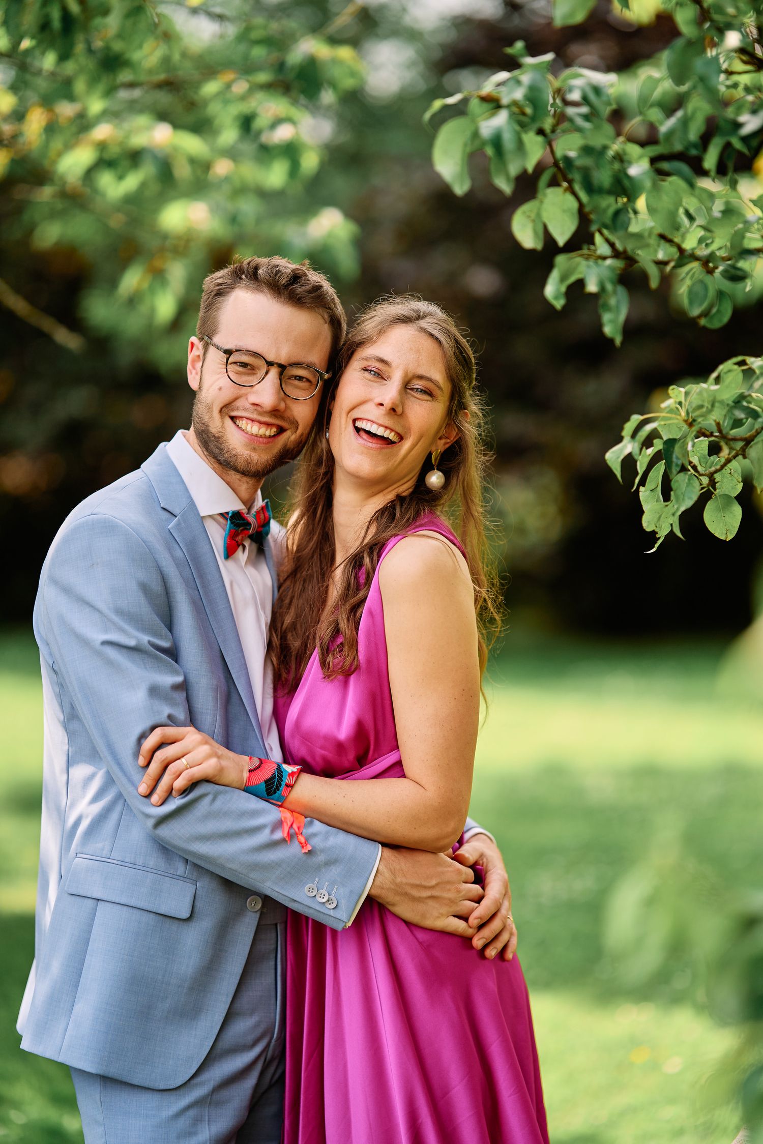 A couple shares joyful laughter together in a garden setting wearing a blue suit and magenta dress.