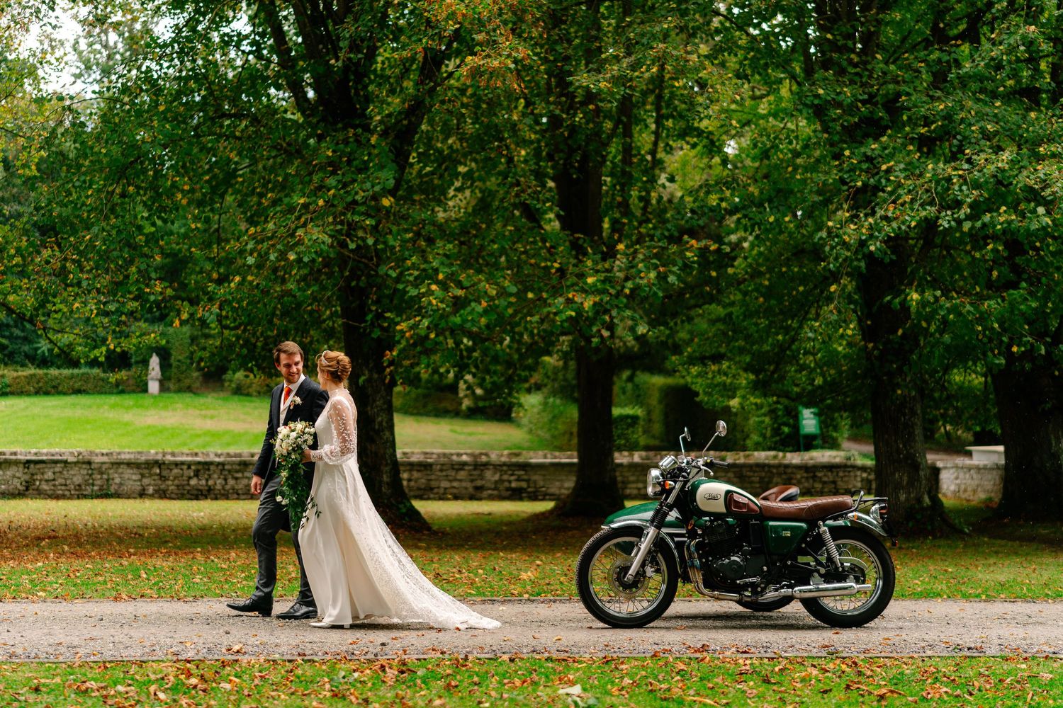 A romantic wedding photo shoot in a lush green park with a vintage motorcycle parked nearby.