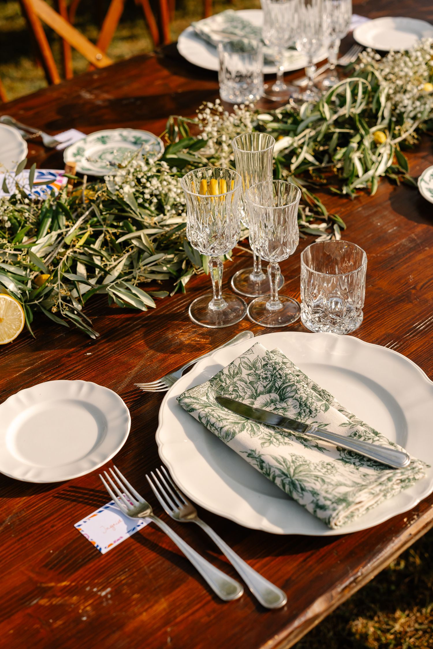Rustic table setting with white plates, crystal glasses, and delicate greenery garland on dark wooden surface.