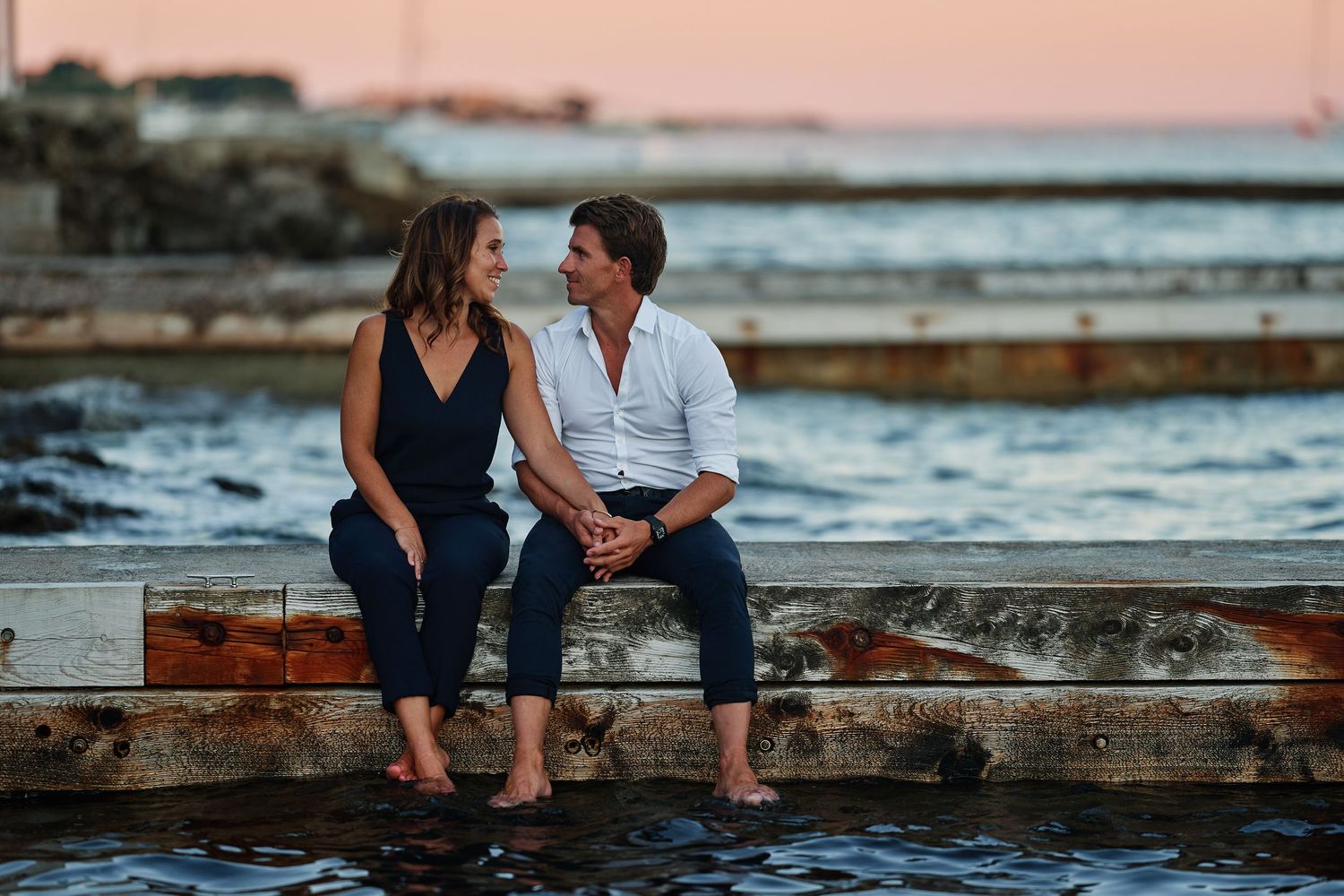 Couple sitting together on stone steps by the ocean during sunset creating a romantic silhouette.
