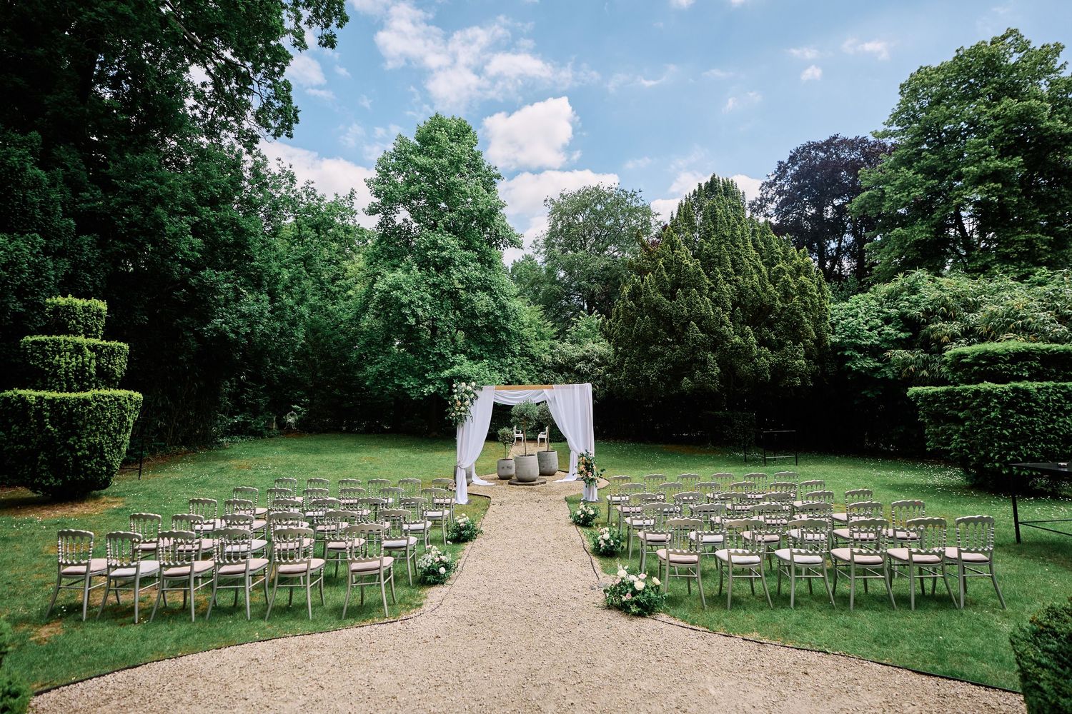 A scenic outdoor wedding ceremony setup with white chairs arranged in rows leading to an elegant white arch in a lush garden setting.