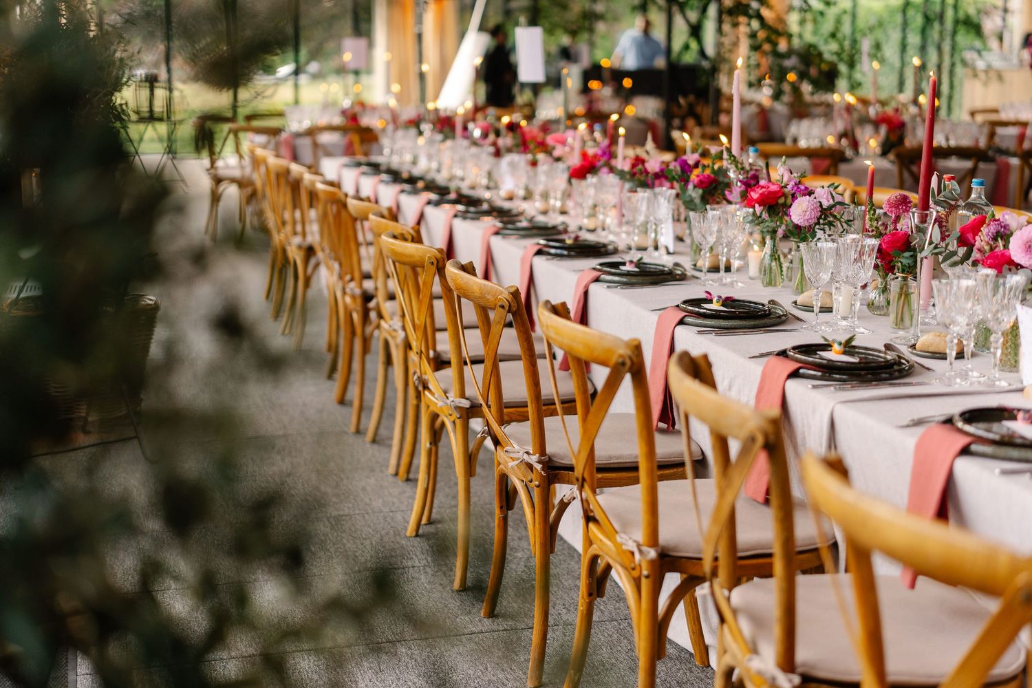 Long banquet tables with elegant place settings and wooden chairs set up for an outdoor wedding reception.