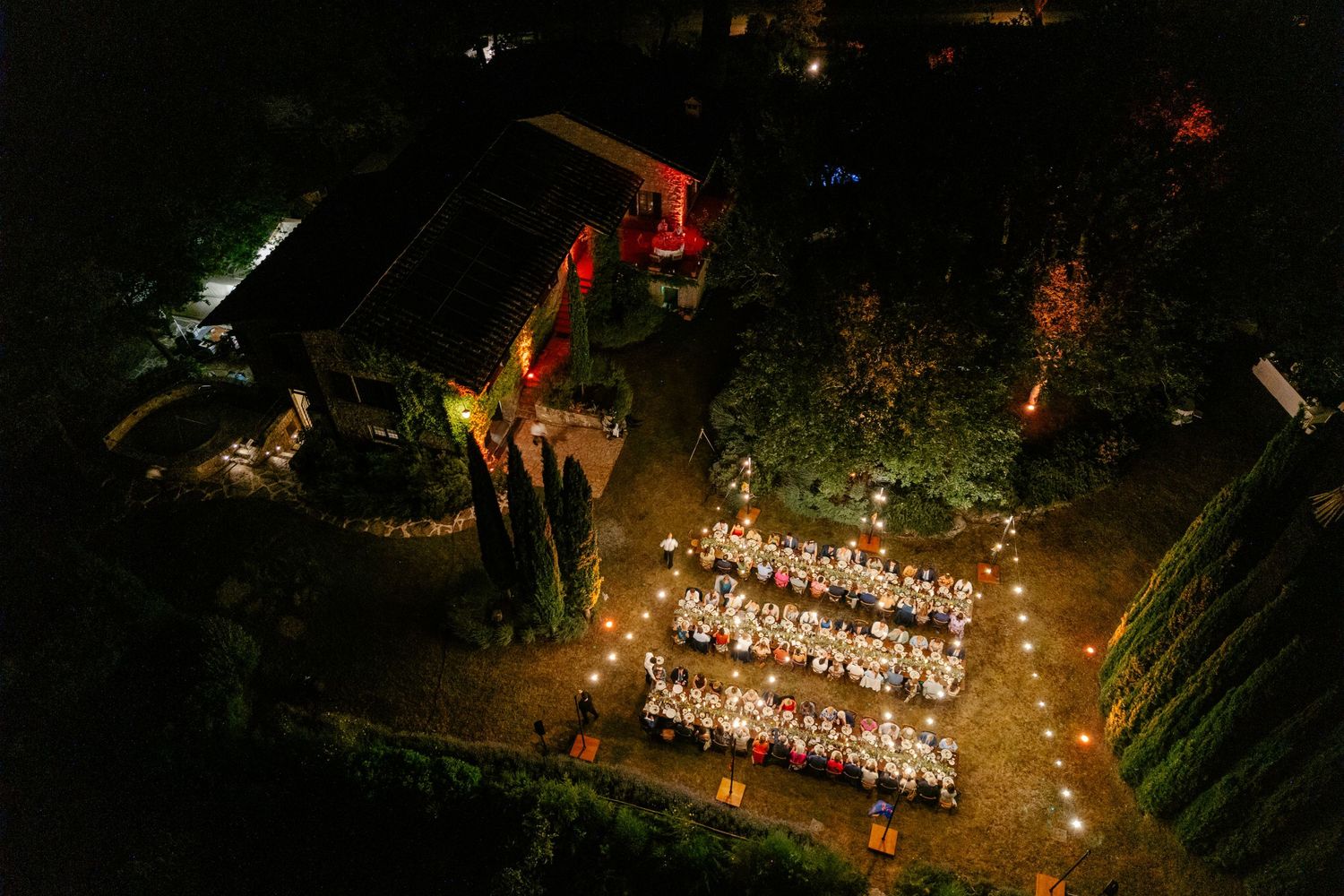 Aerial view of an elegant outdoor wedding reception with long tables illuminated by string lights at night.