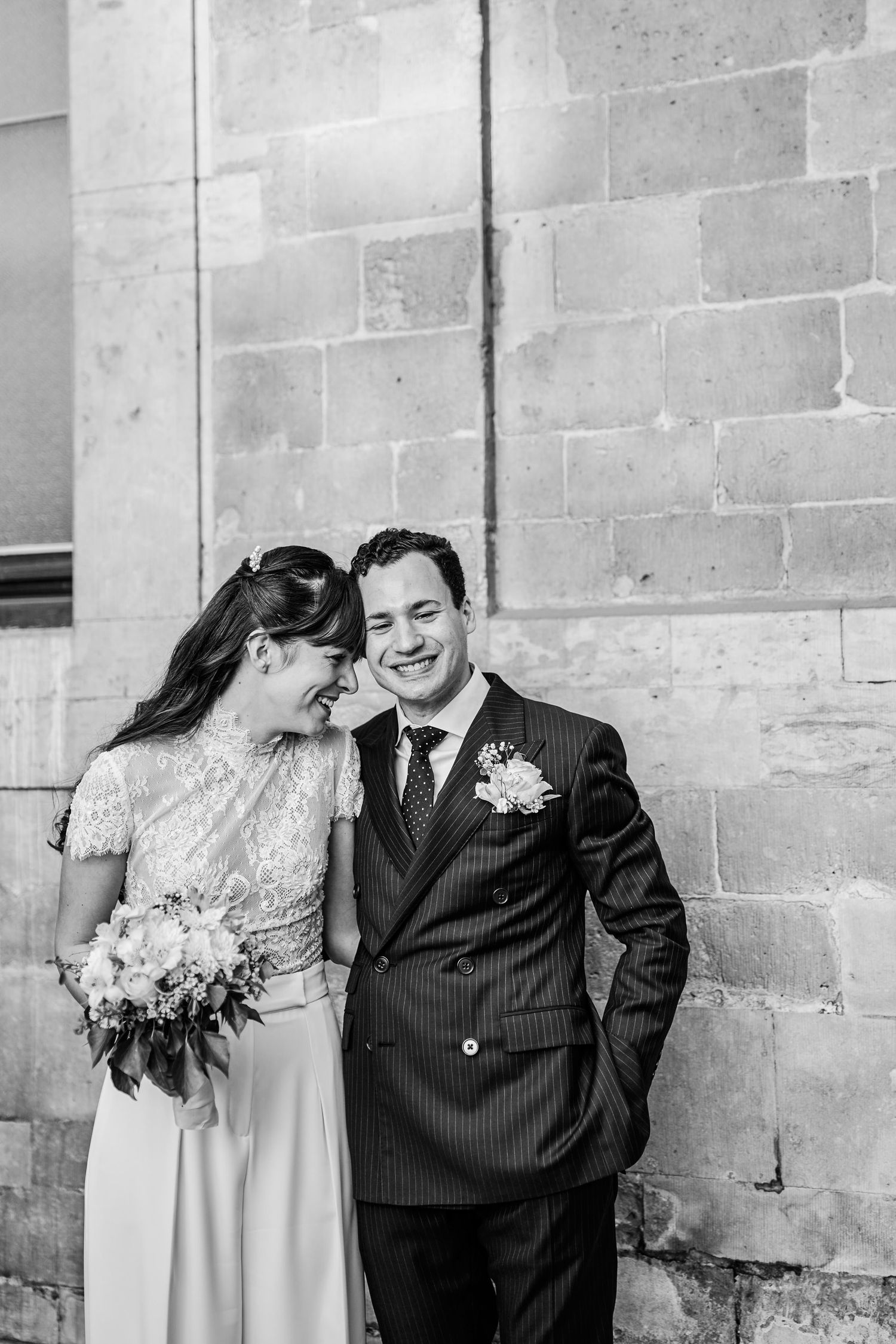 A happy couple shares a joyful moment together against a stone wall during their wedding celebration.