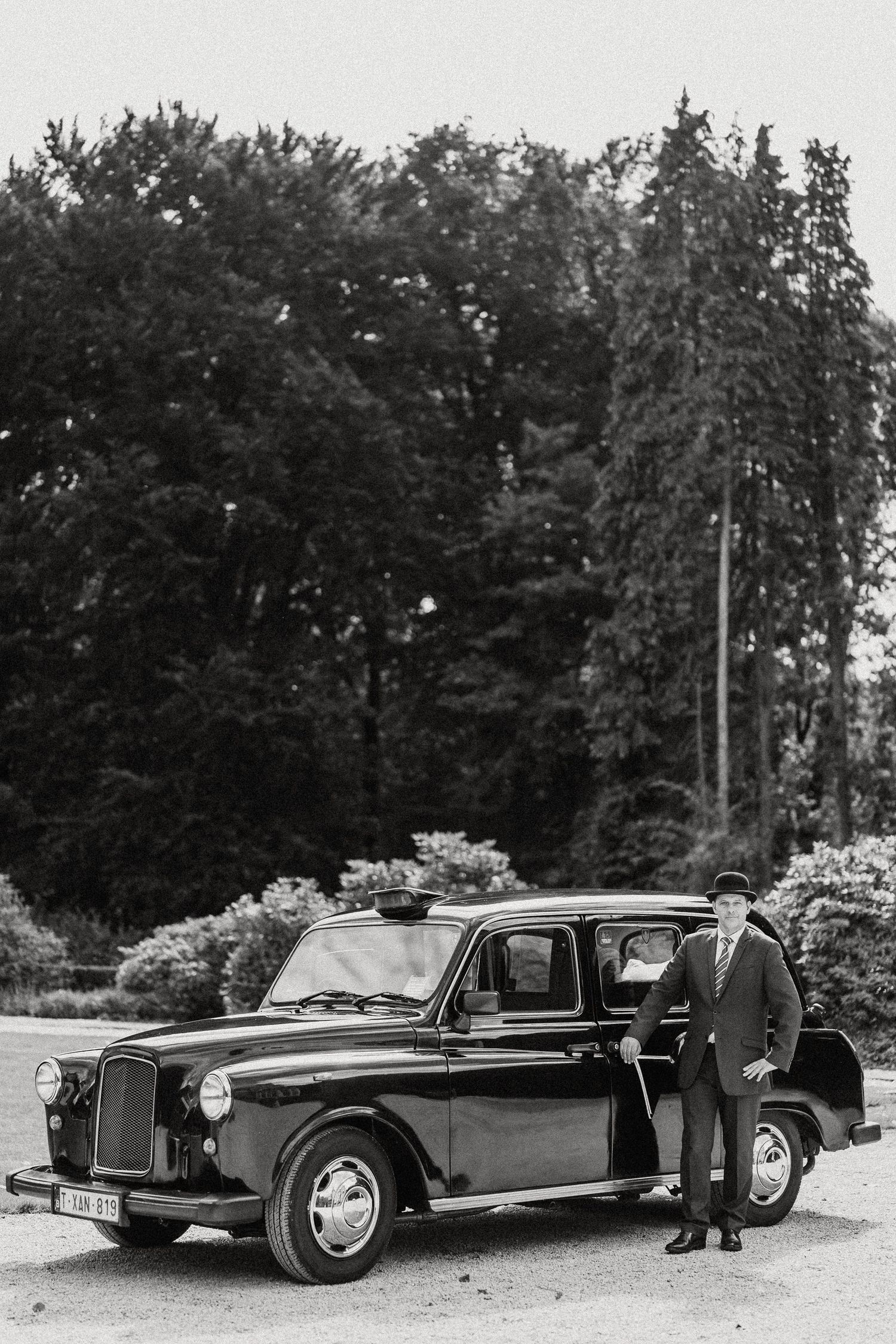 A vintage black Austin taxi car parked in front of trees with someone standing beside it in a black and white photograph.