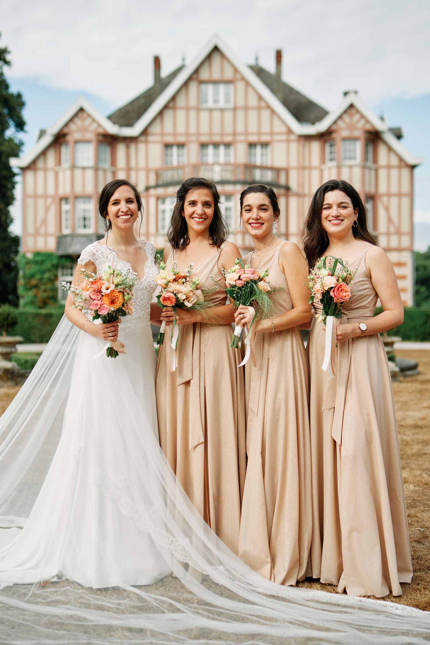 A bridal party in blush-colored bridesmaid dresses and a white wedding gown pose together outside a Tudor-style building.
