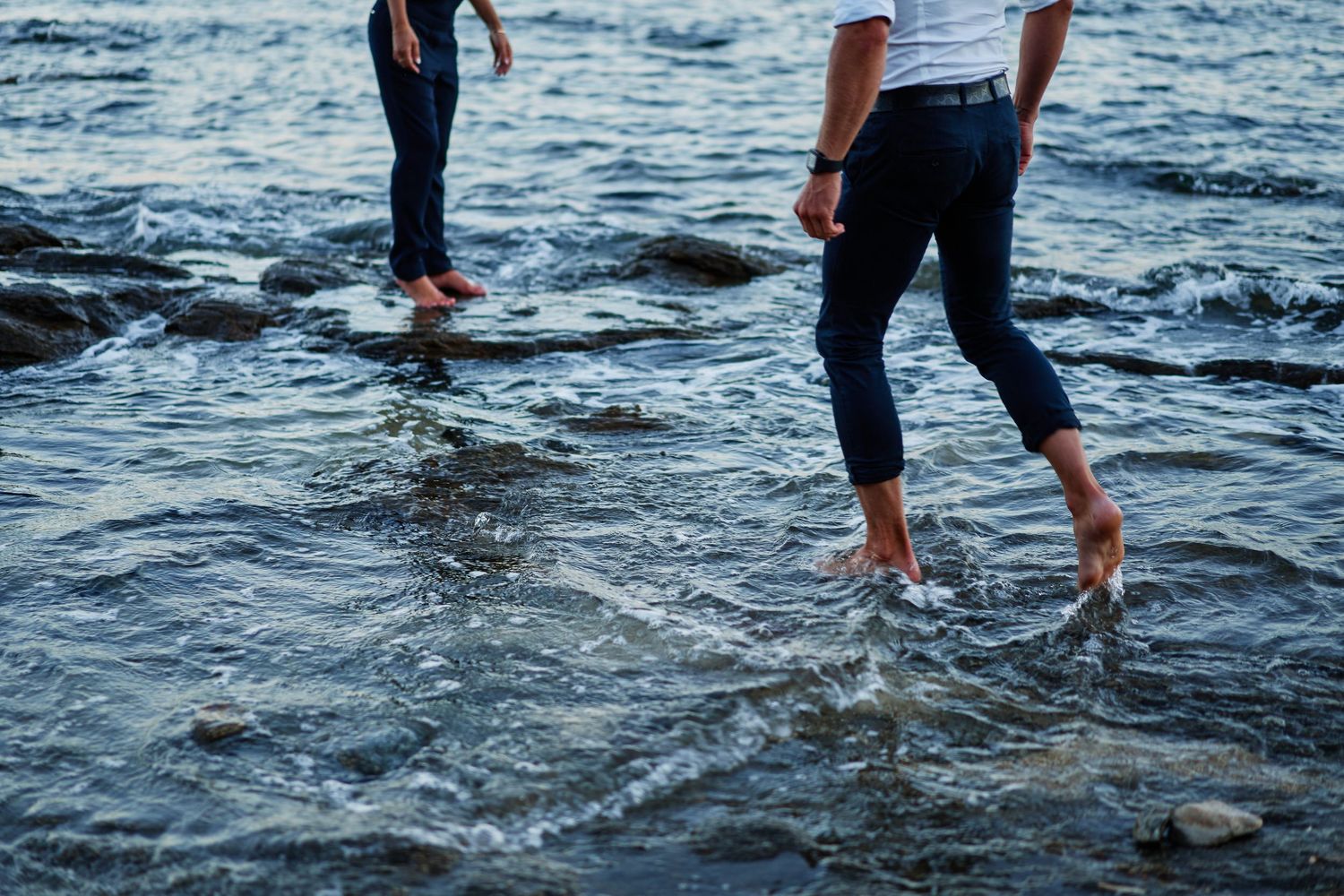 People wading in shallow ocean waves with their feet and legs visible against dark blue rippling water.
