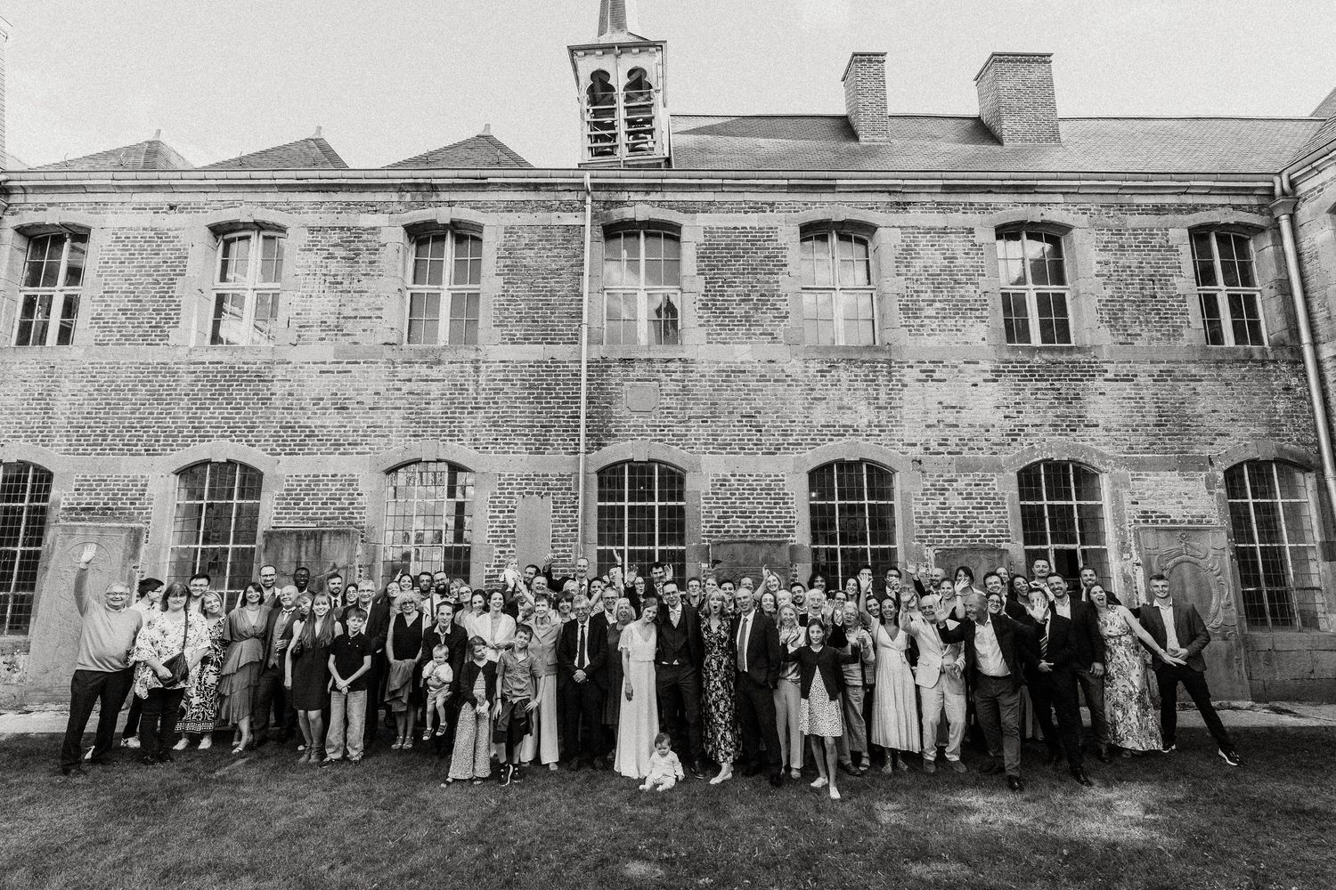 A black and white panoramic photograph shows a large group of people gathered in front of a historic stone building.