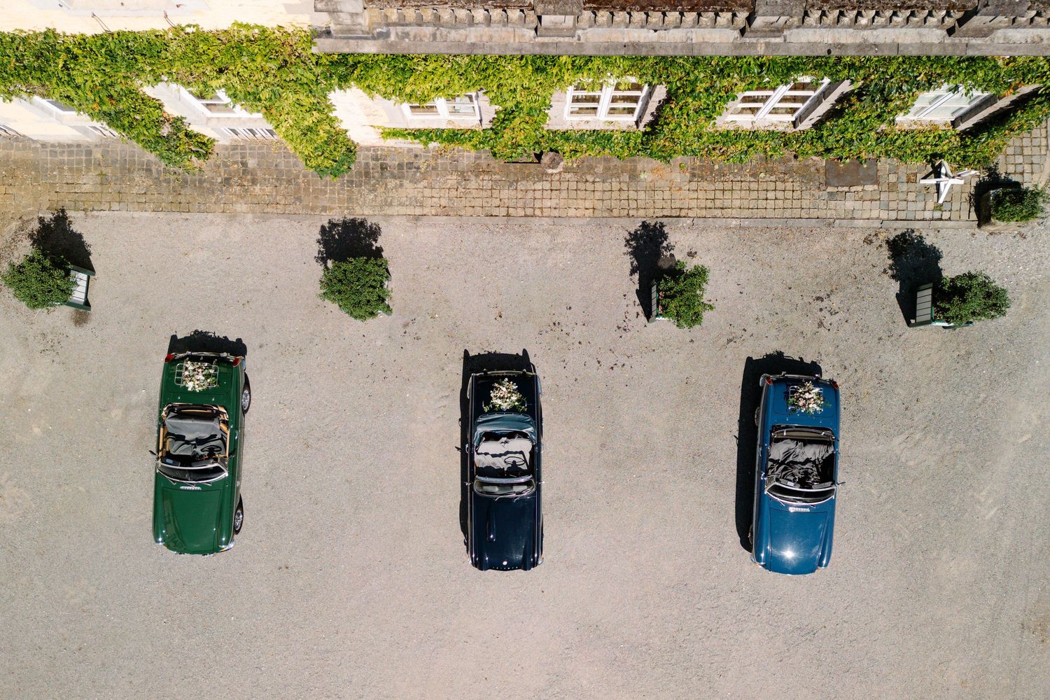 Aerial view of three vintage cars parked in a row on a concrete surface with trees along the side.