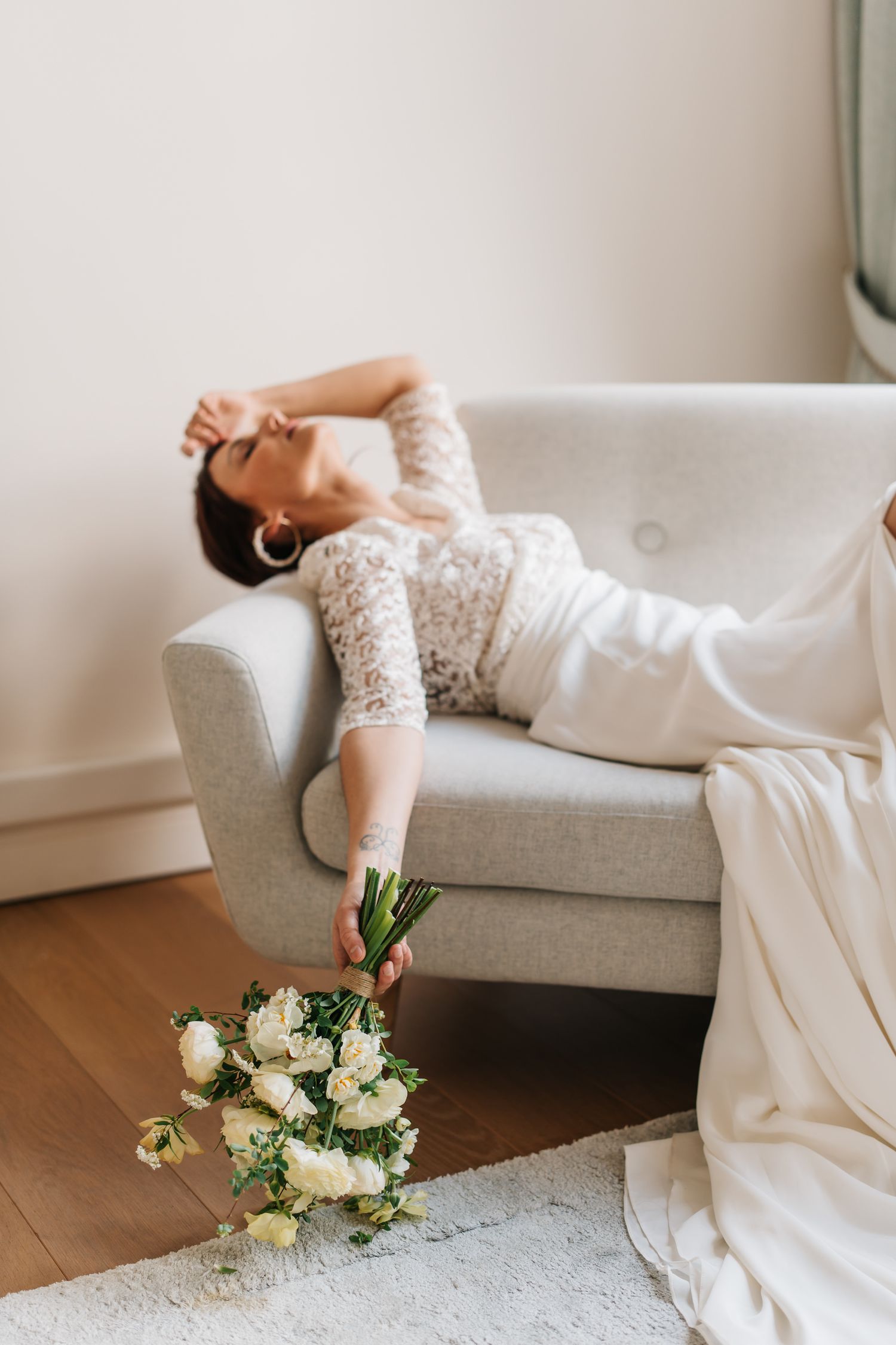 Elegant white lace wedding dress drapes over a modern gray couch with fresh white floral bouquet on floor.