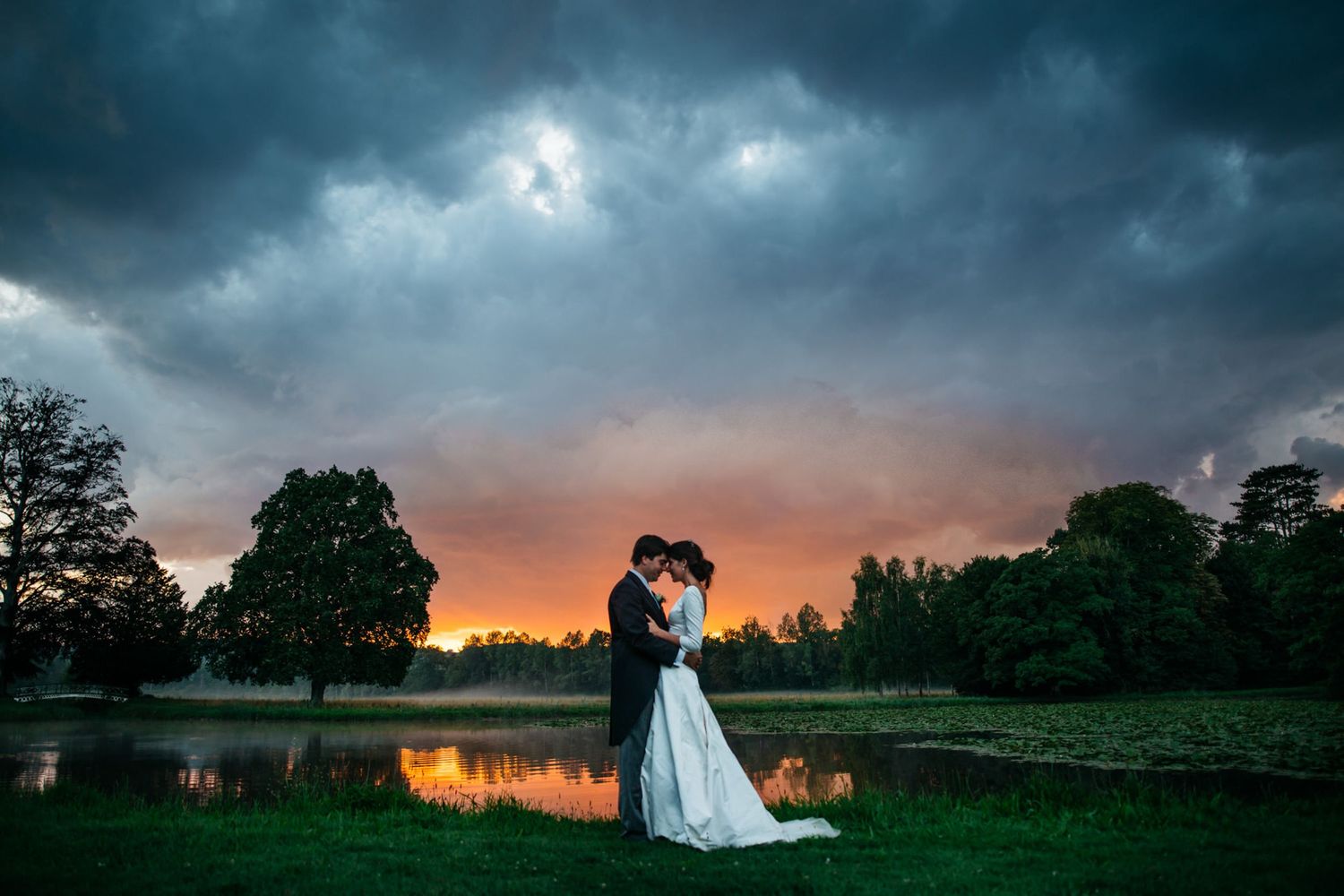 A couple embraces by a lake at sunset with dramatic storm clouds overhead and trees silhouetted against an orange sky.
