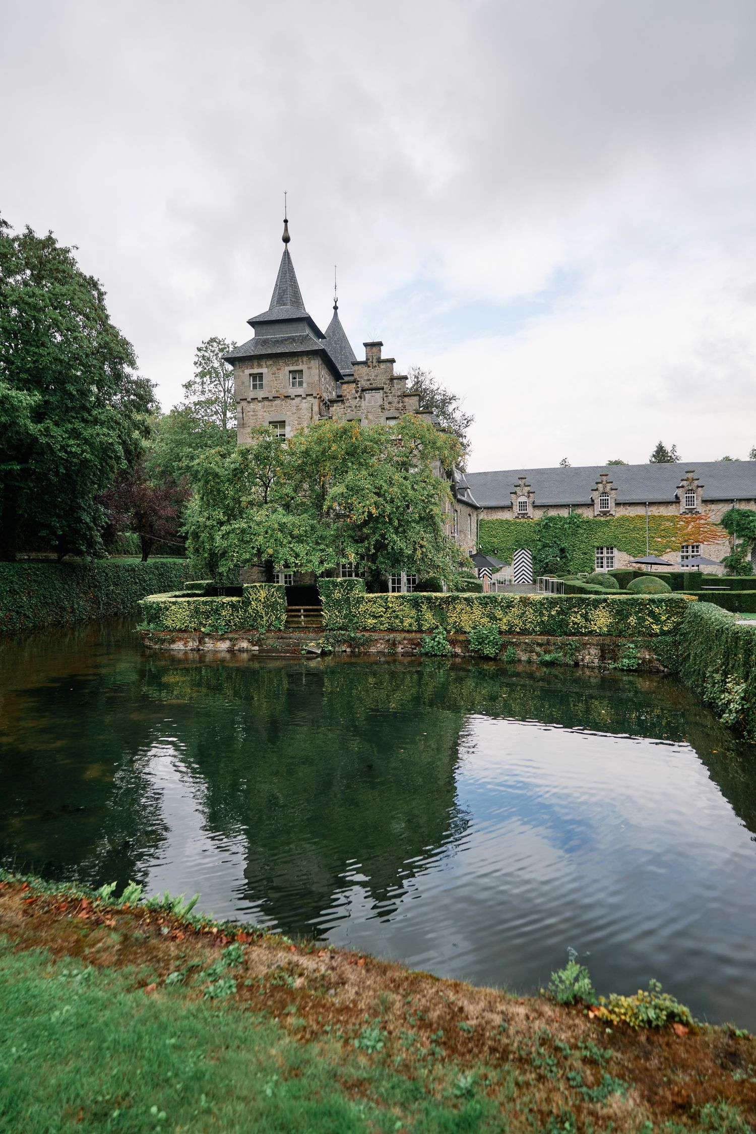 Serene pond with castle turret and spires visible through trees and gardens in a tranquil European estate setting.