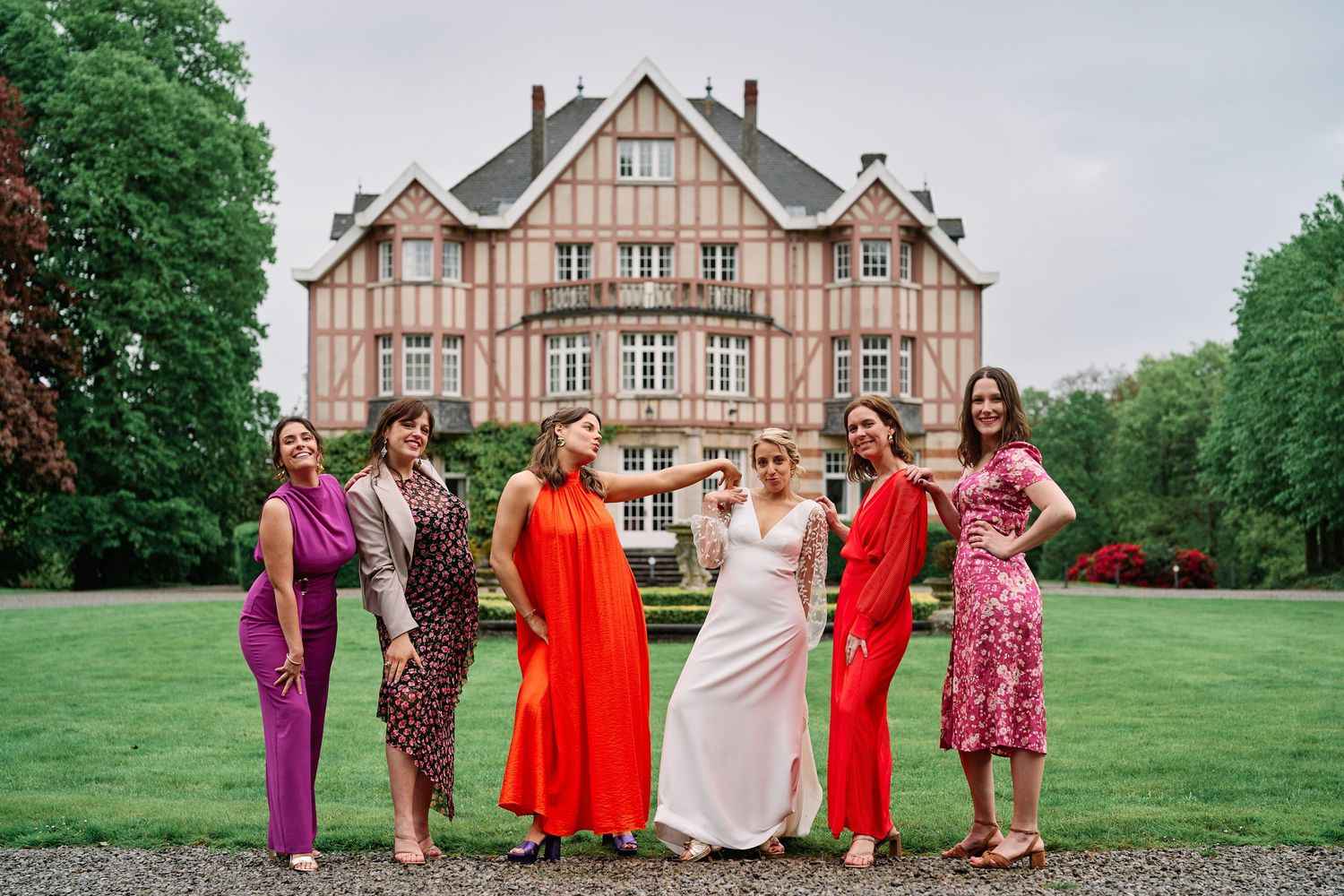 Group in colorful formal dresses pose together on a lush green lawn with a brick mansion in the background.